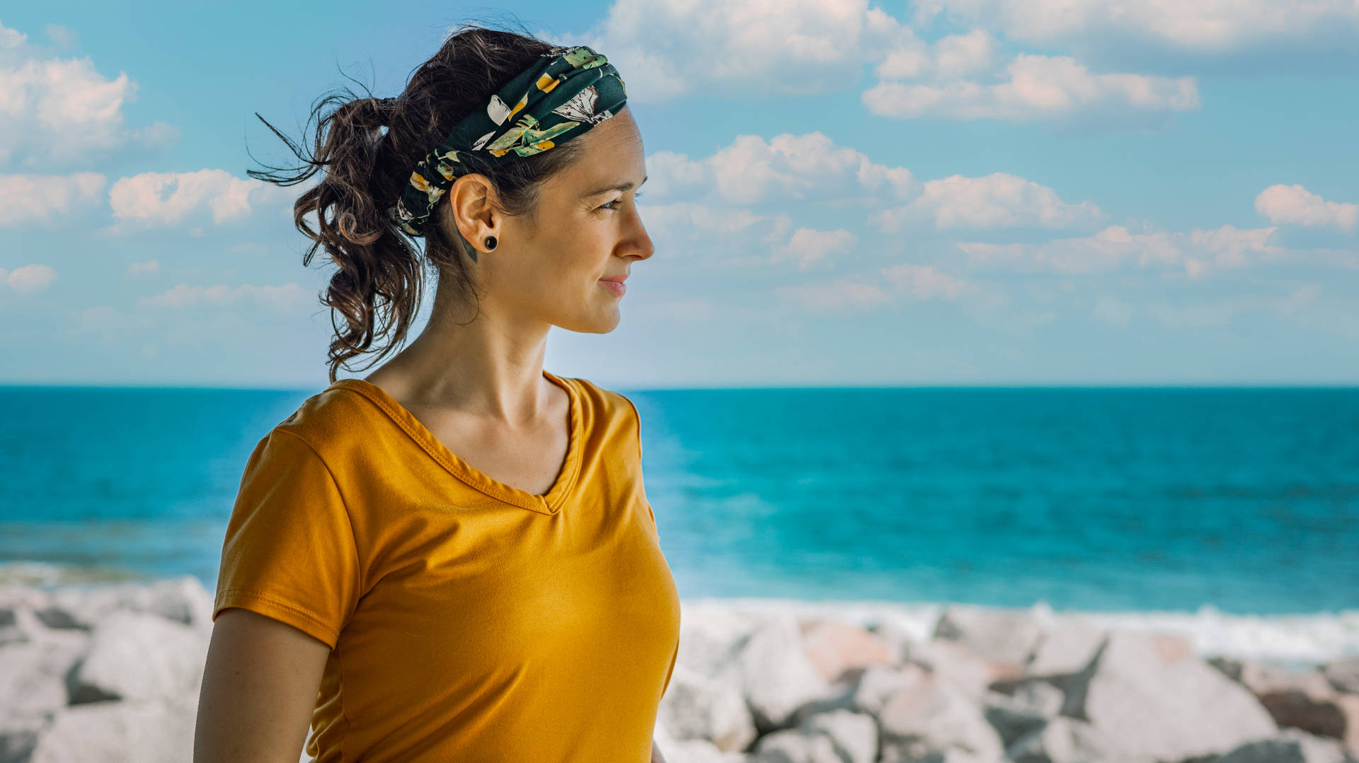 身穿黃色上衣和頭帶的女性站在海邊岩石旁眺望大海。 — 高解析度桌面 壁紙