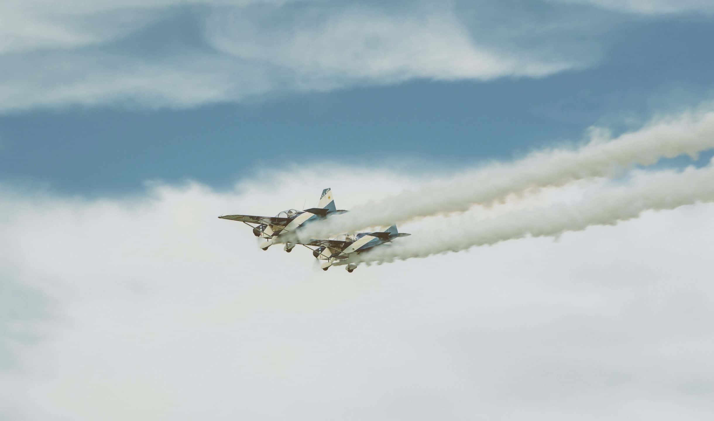 高解析度欣賞小飛機之美 壁紙 — 兩架復古雙翼機編隊飛過多雲的天空，留下濃厚的白色煙霧痕跡。