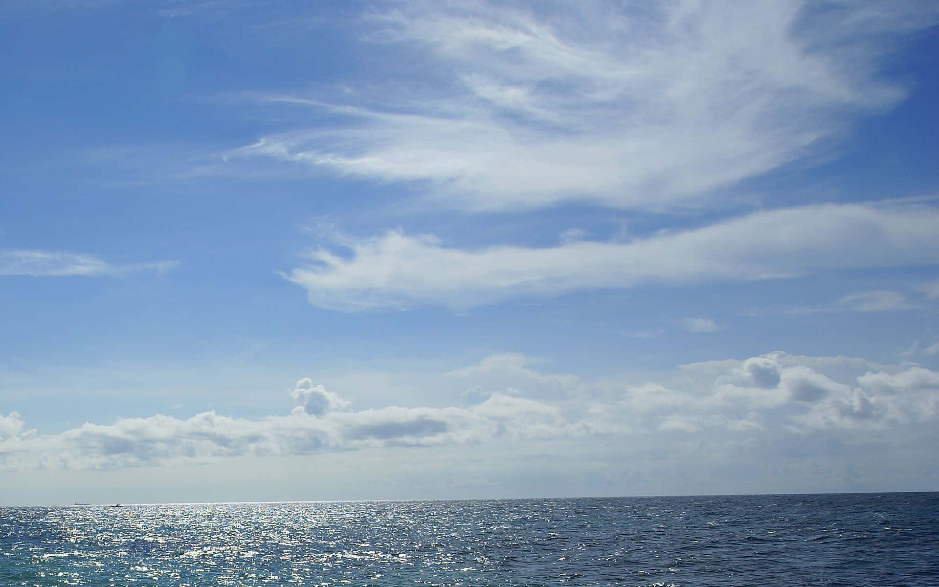 高解析度海邊天空 壁紙 — 廣闊的海洋在明亮的藍天下延伸，點綴著縷縷白雲，水面泛起陽光照射的漣漪。
