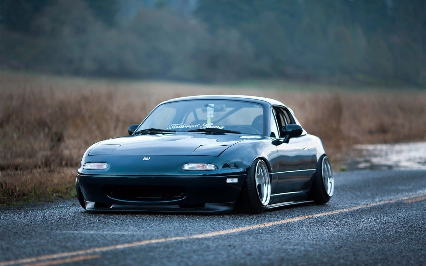 高解析度馬自達MX-5 Miata敞篷車影像 壁紙 — 一架時尚深青綠色Mazda Miata配備訂製輪圈，低趴於碎石路上，旁邊是乾草，沐浴在柔和暮光之中。