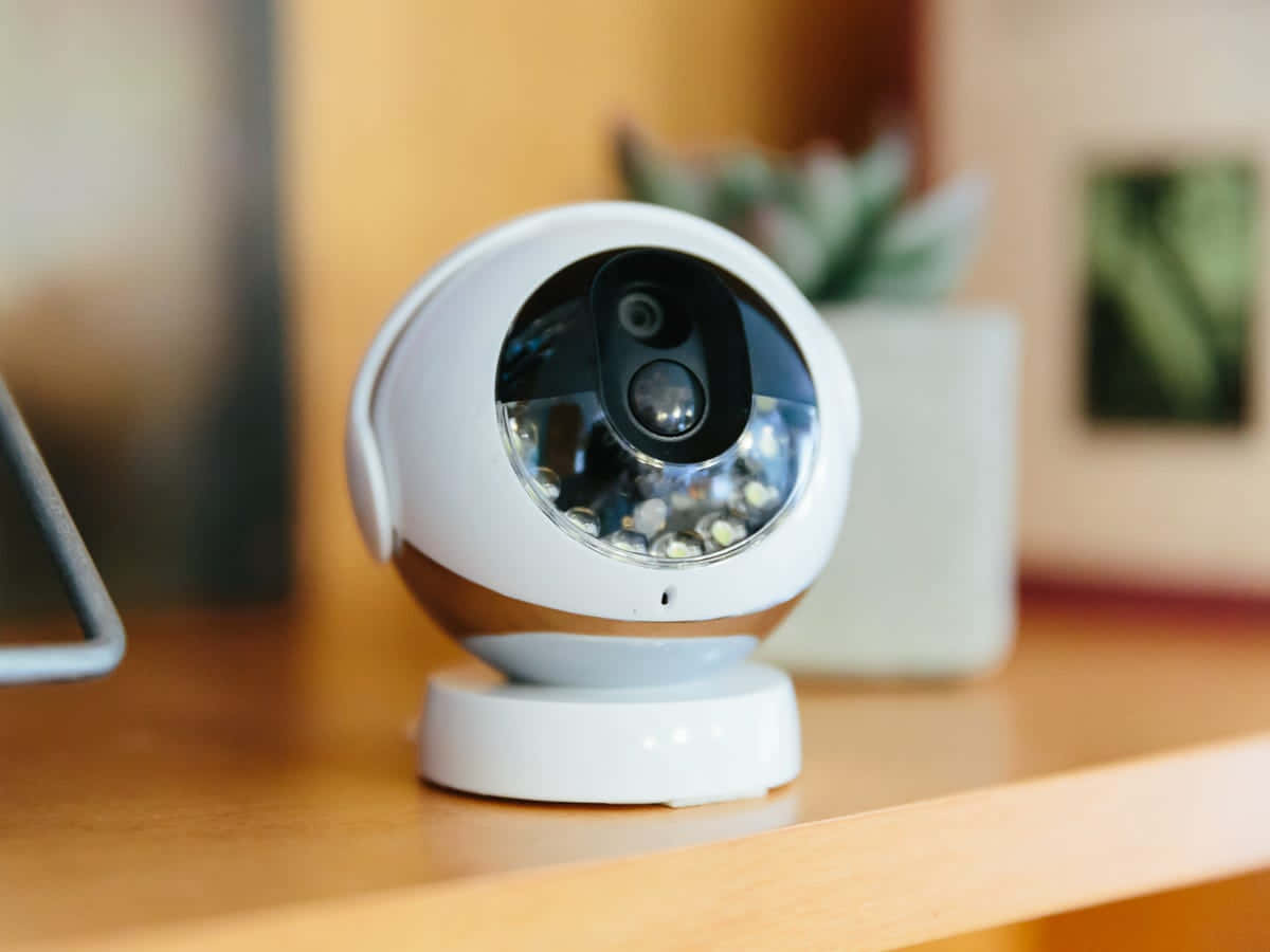 高階 Kidde Remotelync 無線安全攝影機 壁紙 — 一部時尚的白色安全攝影機放置在木架上，背景是柔和的散景效果，包括室內植物和裱框藝術品。
