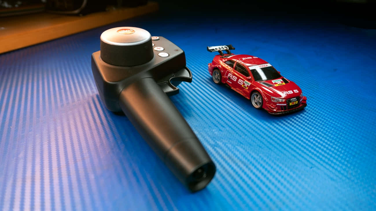 高飛RC車，為您帶來刺激與冒險 壁紙 — 一個黑色賽車控制器和紅色Audi RS5玩具車放在藍色紋理表面上，在聚焦燈光下。