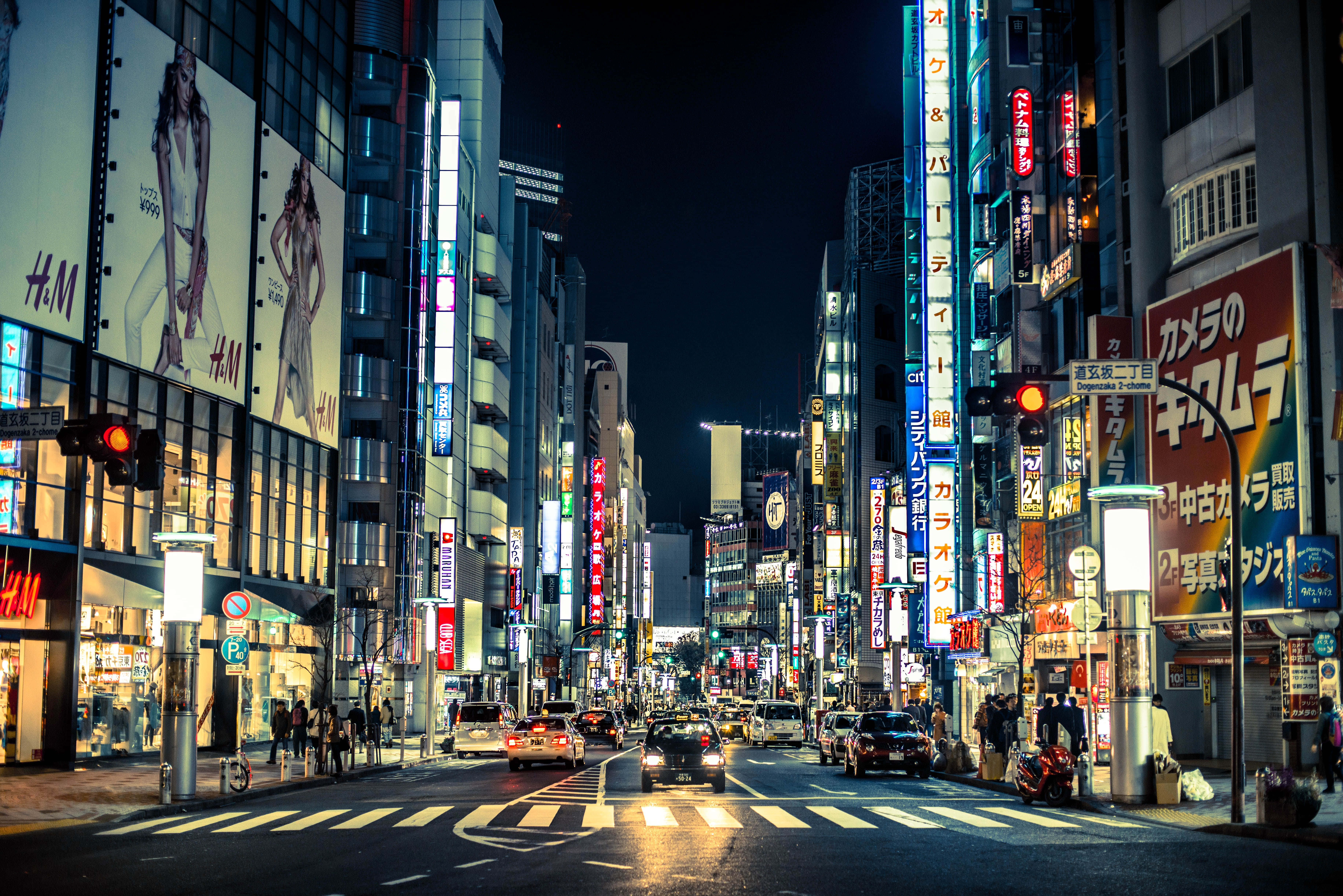 高高翱翔於繁忙的卡通城上空 壁紙 — 充滿活力的東京夜晚街道，霓虹燈閃爍，發光的招牌、汽車和熙攘的行人穿梭在高聳建築物之間。