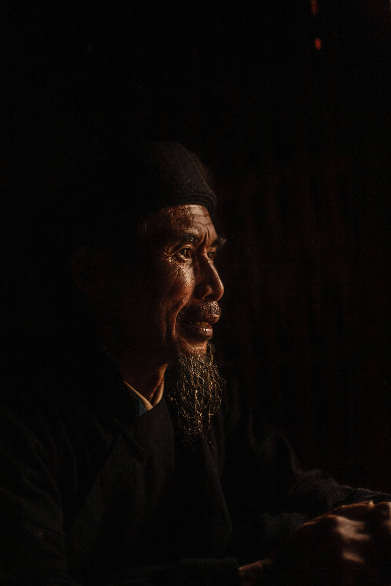 一位留著長鬍鬚的老人，在昏暗的燈光下若有所思地望向遠方。