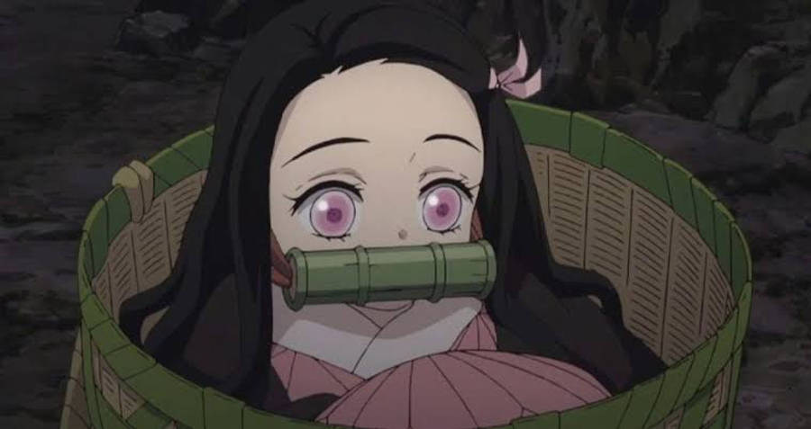 鬼滅之刃 祢豆子 壁紙 — 一位年輕的動漫女孩，粉紅色眼睛和黑色頭髮，坐在編織籃子裡，嘴巴被竹管覆蓋，在黑暗的岩石環境中。