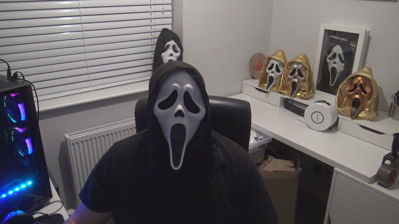 戴著Ghostface面具的人坐在電腦桌前，周圍有恐怖收藏品。