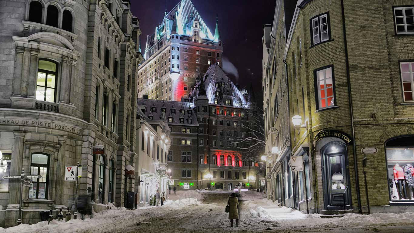 魁北克市前衛城堡雪景 壁紙 — 夜晚，一個孤獨的身影走在積雪的鵝卵石街道上，朝著魁北克市燈火通明的芳堤娜城堡走去。