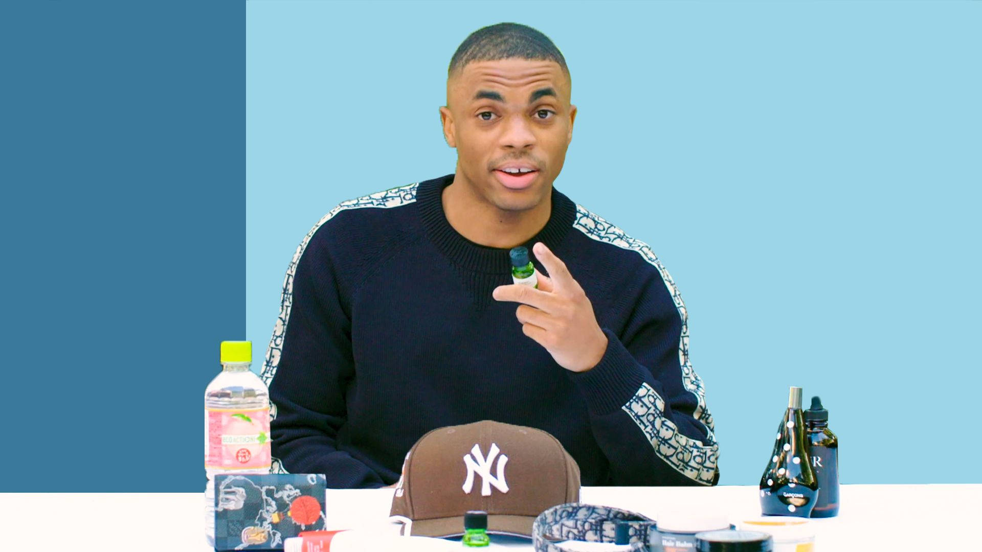魅力 Vince Staples 壁紙 — 一個穿著黑色圖案袖毛衣的男人坐在桌子旁，手持一個小瓶子，背景是雙色調的藍色。