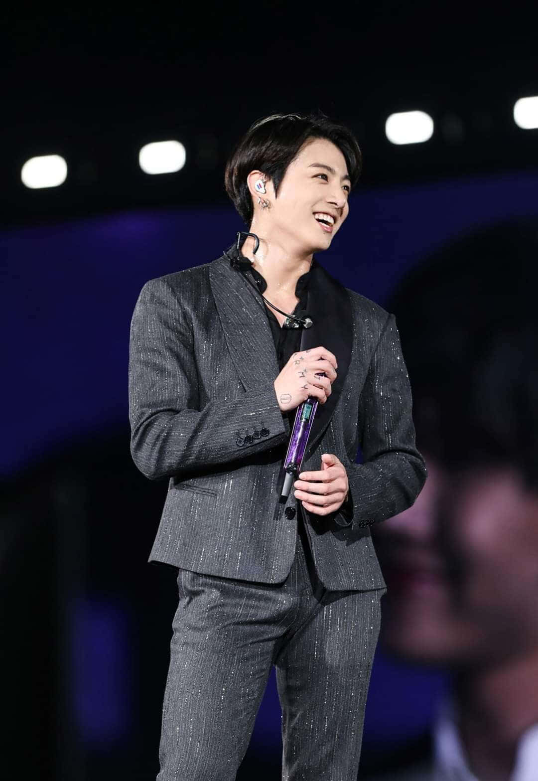 魅力四射的Jungkook時尚造型 壁紙 — 一位微笑的表演者穿著閃亮的灰色西裝，在黑暗背景下明亮的舞台燈光下握著麥克風。