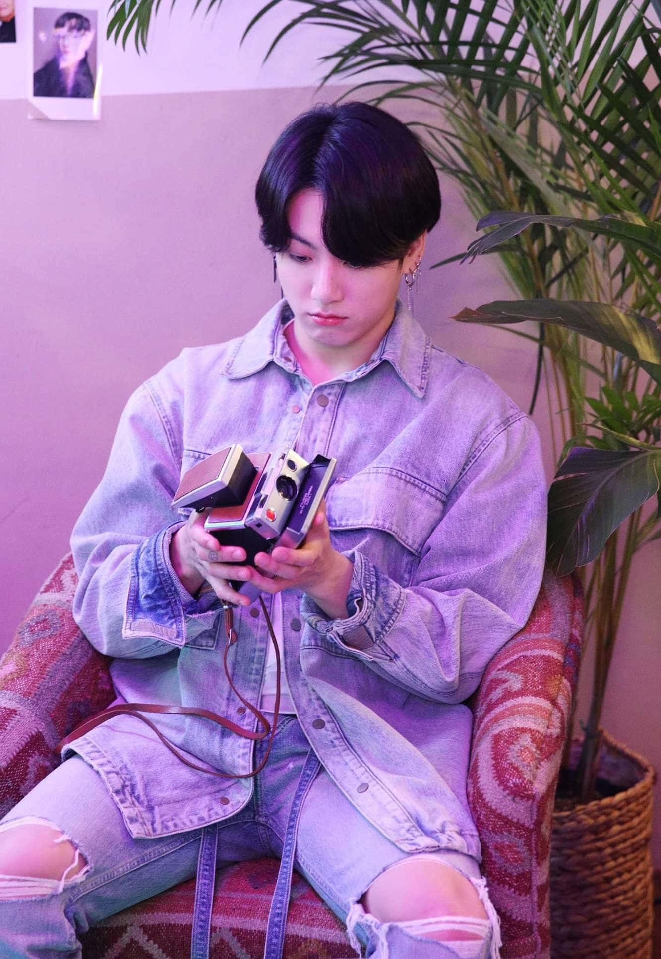 魅力四射的Jungkook肖像 壁紙 — 一位身穿牛仔外套的年輕男子坐在圖案椅子上，在紫色燈光下拿著復古相機，旁邊有一盆盆栽。
