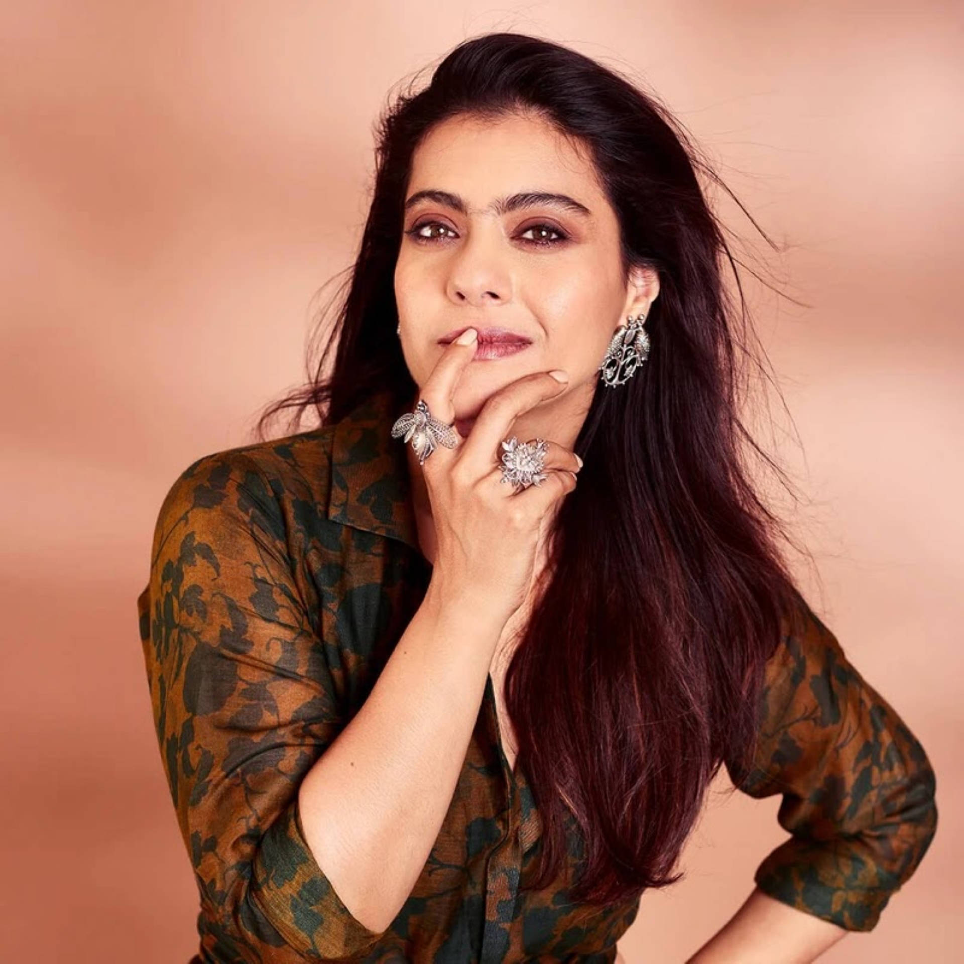 魅力女星 Kajol 壁紙 — 一位深色頭髮、佩戴大膽珠寶的優雅女士，在柔和蜜桃色背景下若有所思地擺姿勢。