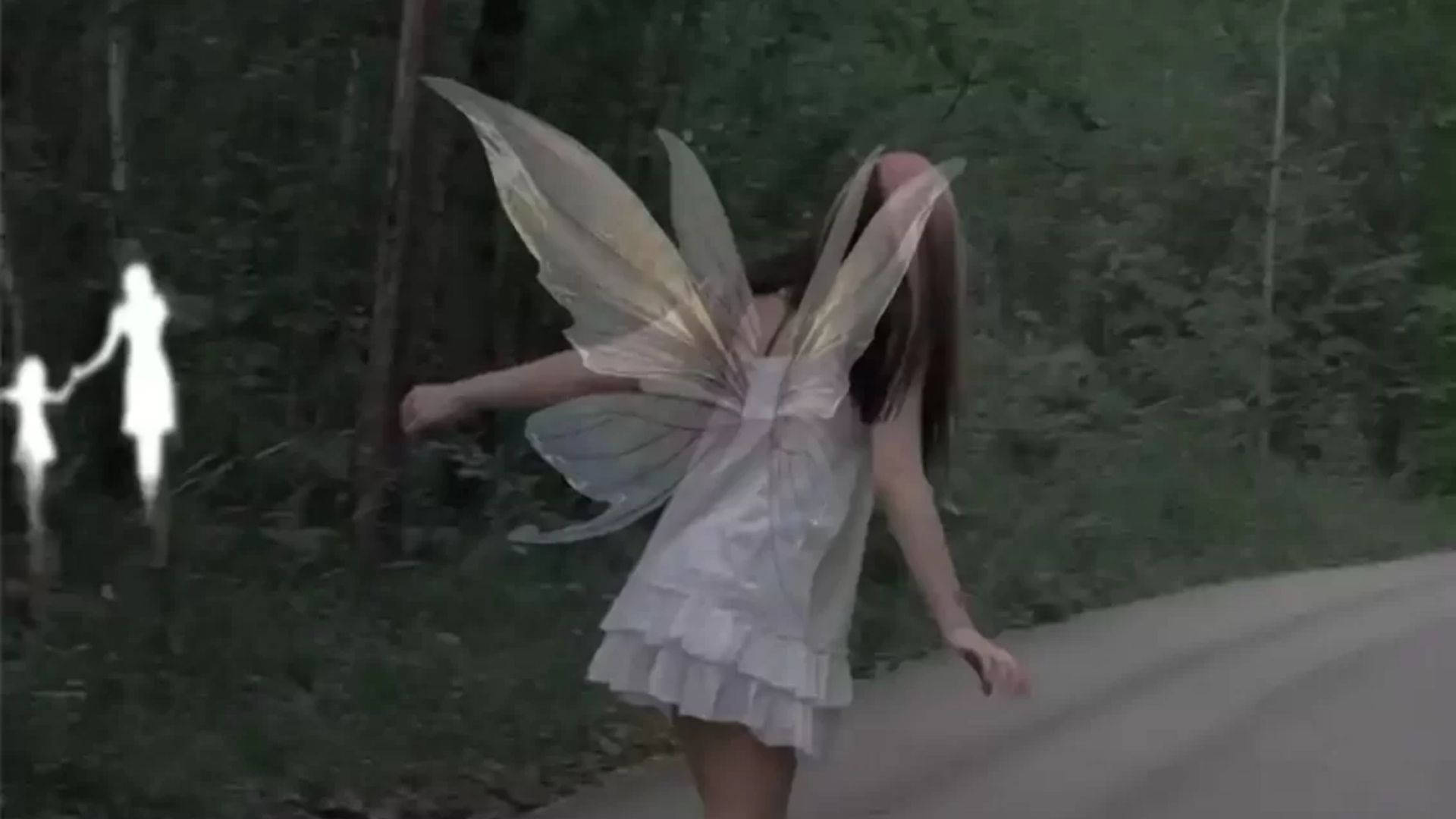 一個穿著白色連衣裙的仙子走在森林道路上，附近有發光的鬼魂般的身影。