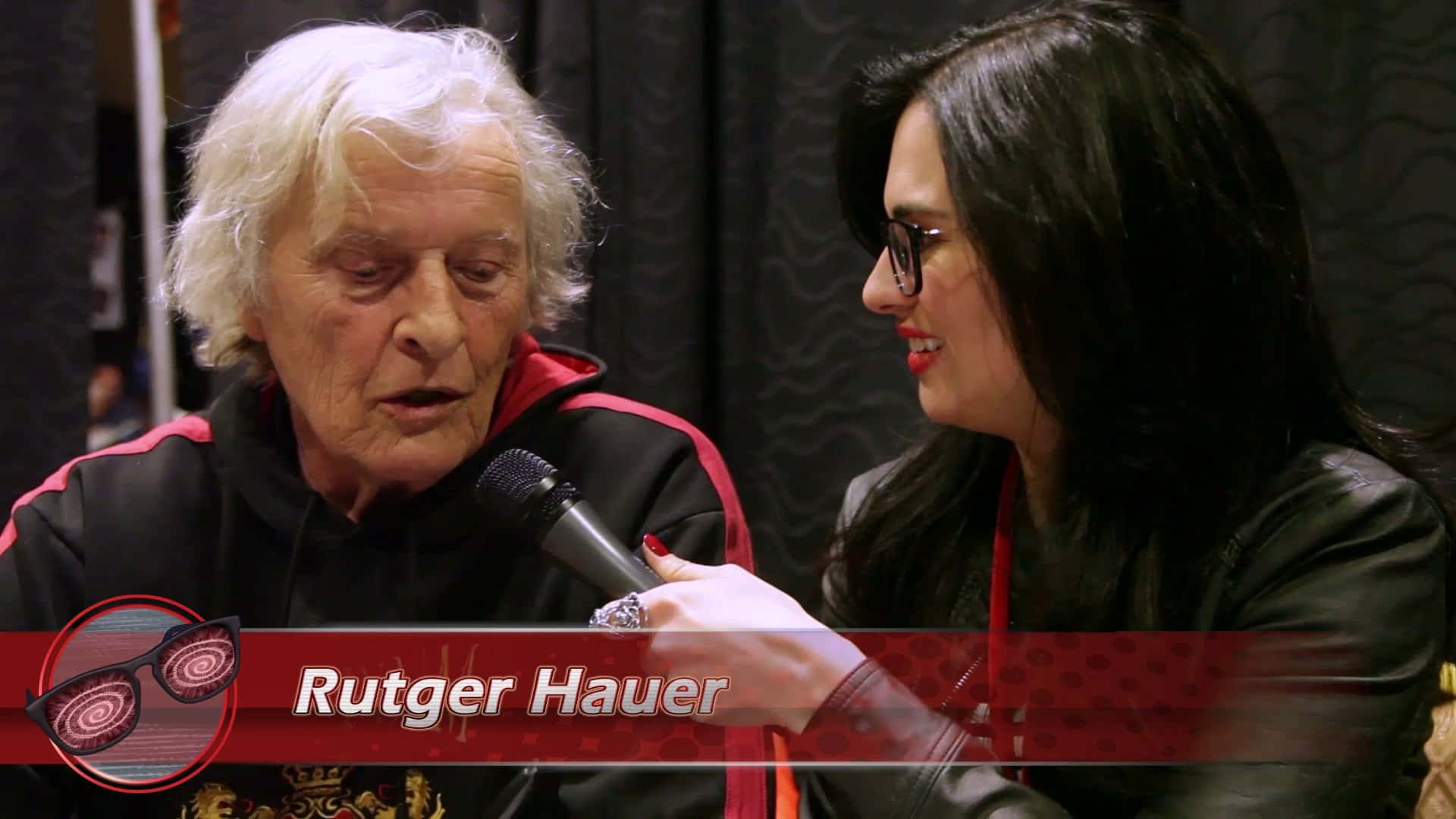 Rutger Hauer 正接受一位戴眼鏡、手持咪高峰的女子訪問。