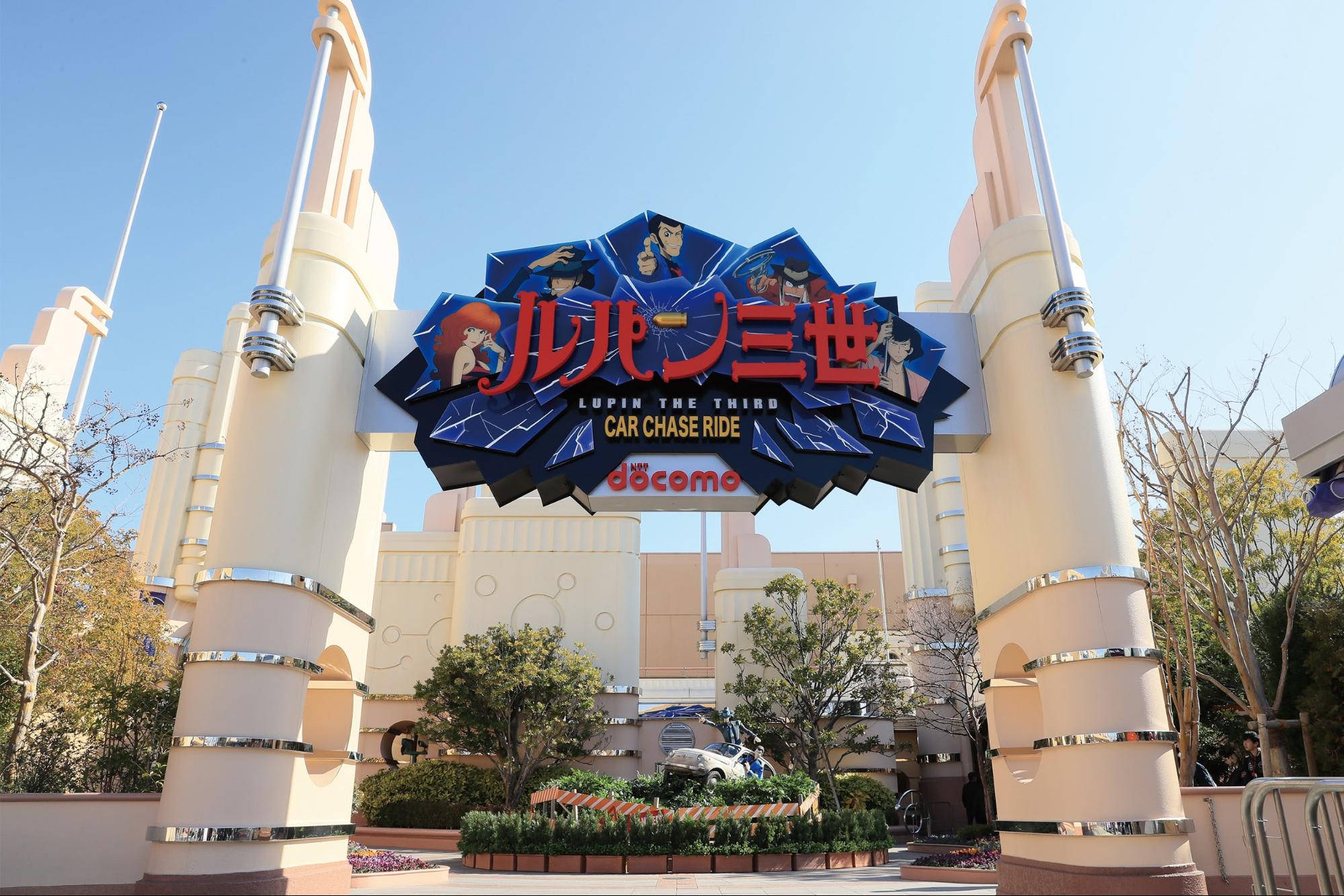 《魯邦三世》環球影城 壁紙 — 明亮的藍天襯托著主題公園中《魯邦三世》汽車追逐之旅的宏偉入口，配有醒目的標誌和奶油色柱子。