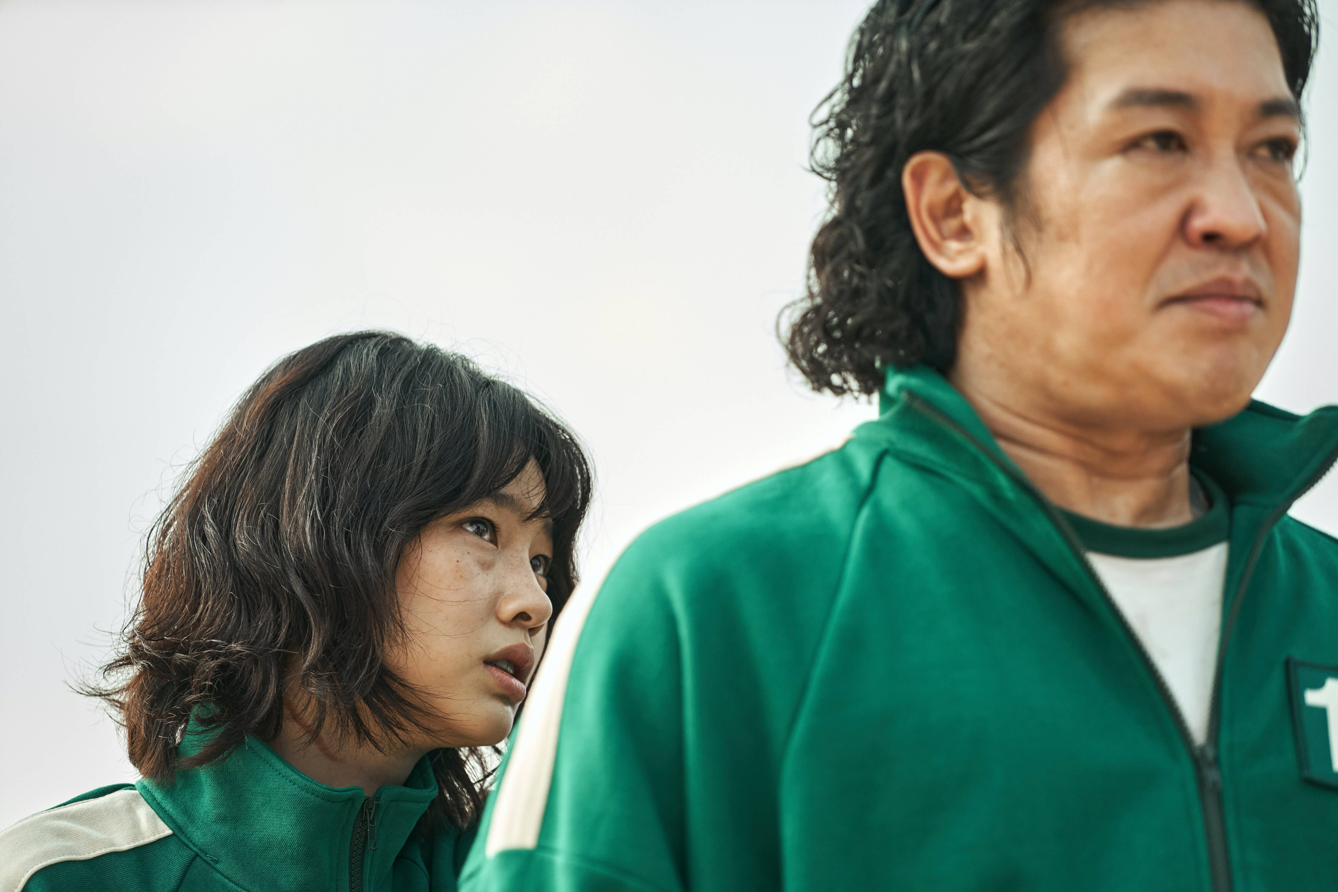 魷魚遊戲 067 壞人背後 壁紙 — 兩個身穿綠色號碼運動服的人，在蒼白的天空下凝視著。