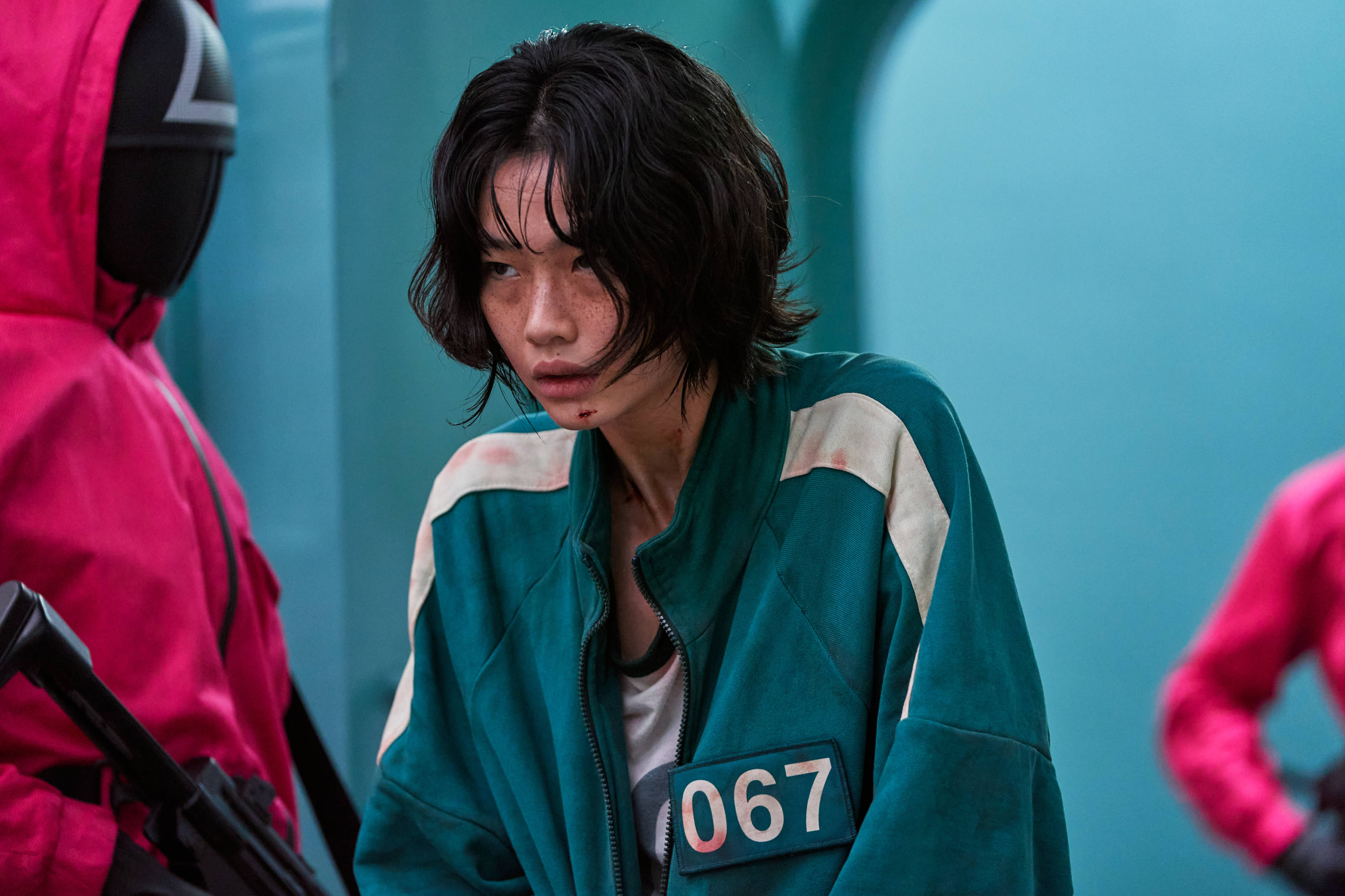 魷魚遊戲 067 新招募 壁紙 — 一位身穿青綠色號碼外套的疲憊參賽者，在緊張的遊戲場景中，對著青綠色的牆壁露出疲憊的眼神。