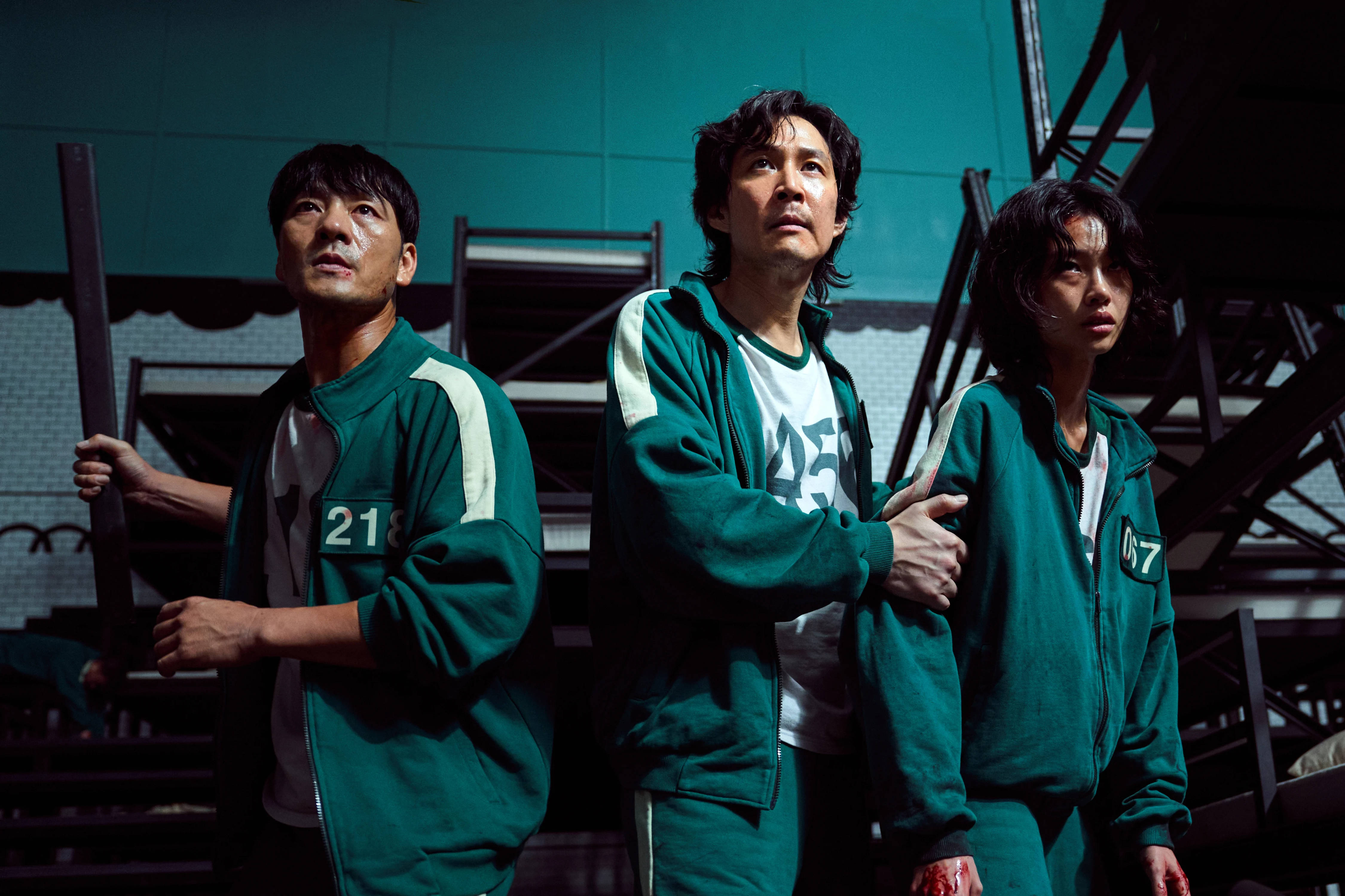 魷魚遊戲 067 最後三人 壁紙 — 三名身穿綠色號碼運動服的緊張參賽者站在一個昏暗的工業空間，內有金屬樓梯和藍綠色牆壁。