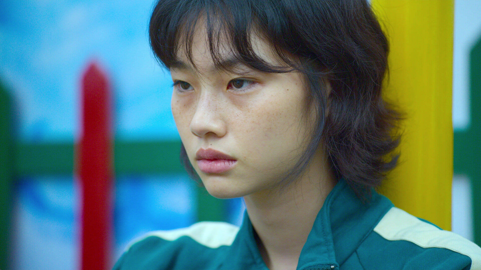 魷魚遊戲 067 糖果遊戲 壁紙 — 一位身穿綠色和白色運動服的年輕女性，在色彩繽紛的模糊背景下若有所思地凝視著。