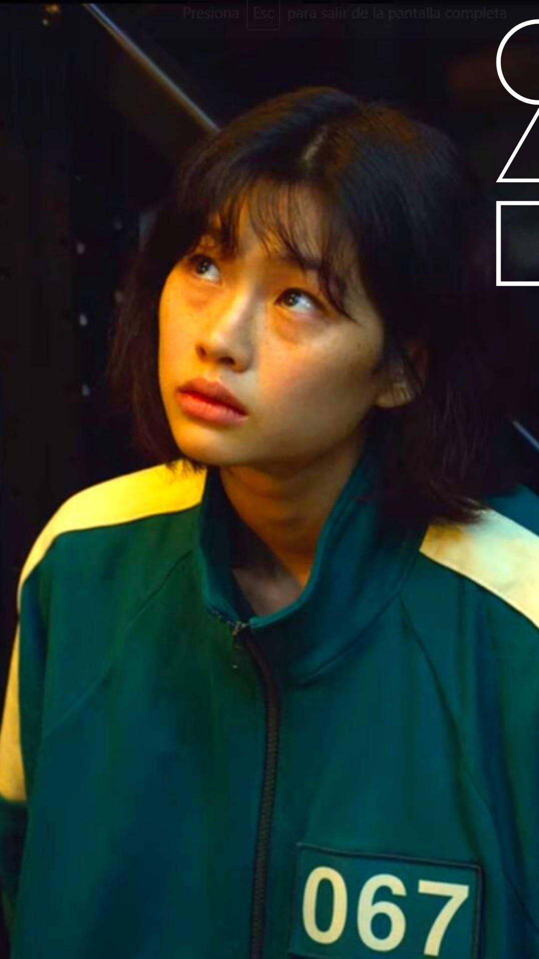魷魚遊戲 067 號仰望 壁紙 — 一位年輕女性身穿綠色和黃色帶號碼的夾克，在昏暗的環境中帶著擔憂的表情向上看。
