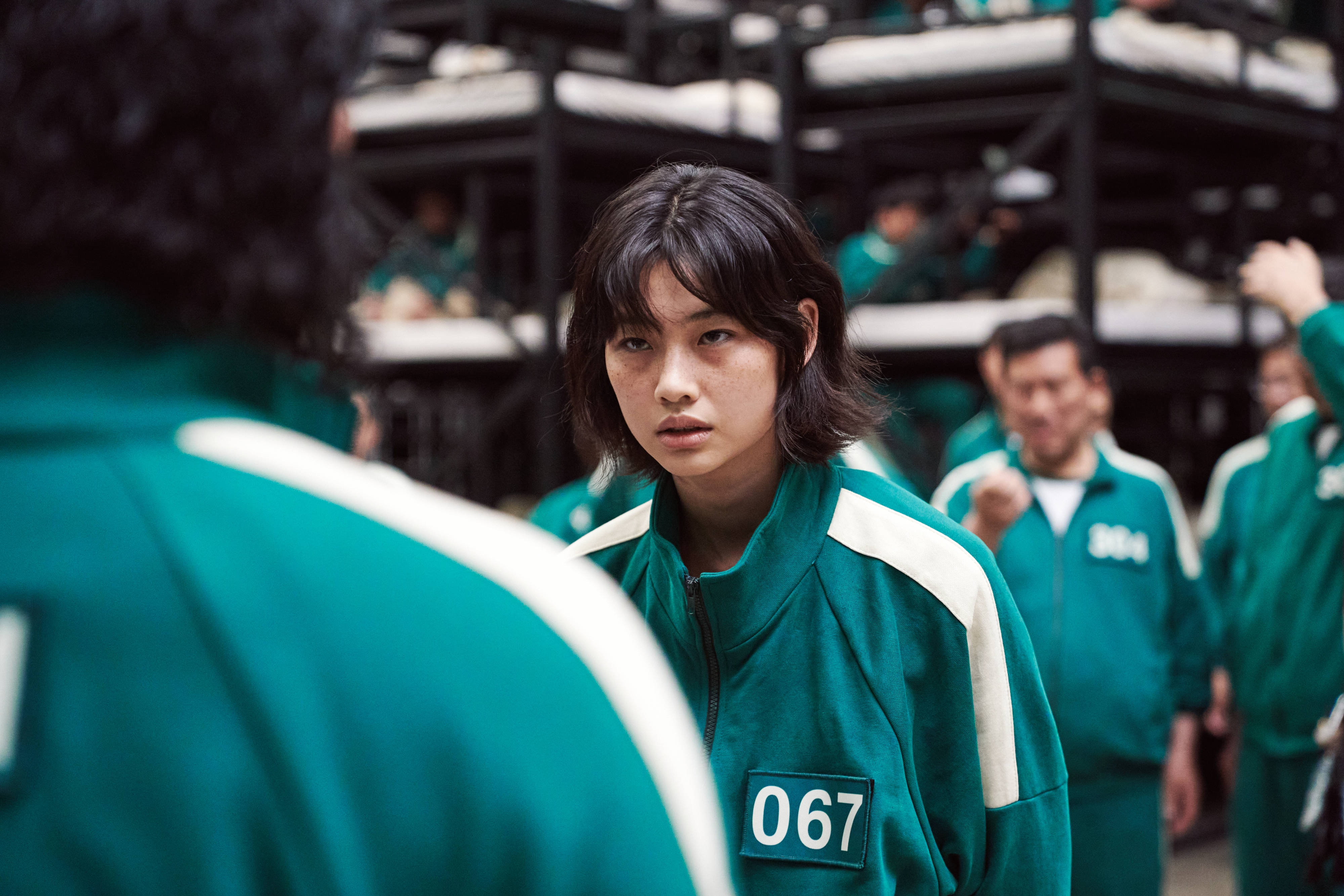 魷魚遊戲 067 號寢室對峙 壁紙 — 一位身穿綠色號碼運動服的年輕女子，在擁擠且燈光昏暗的遊戲競技場中，專注地凝視著另一位玩家。