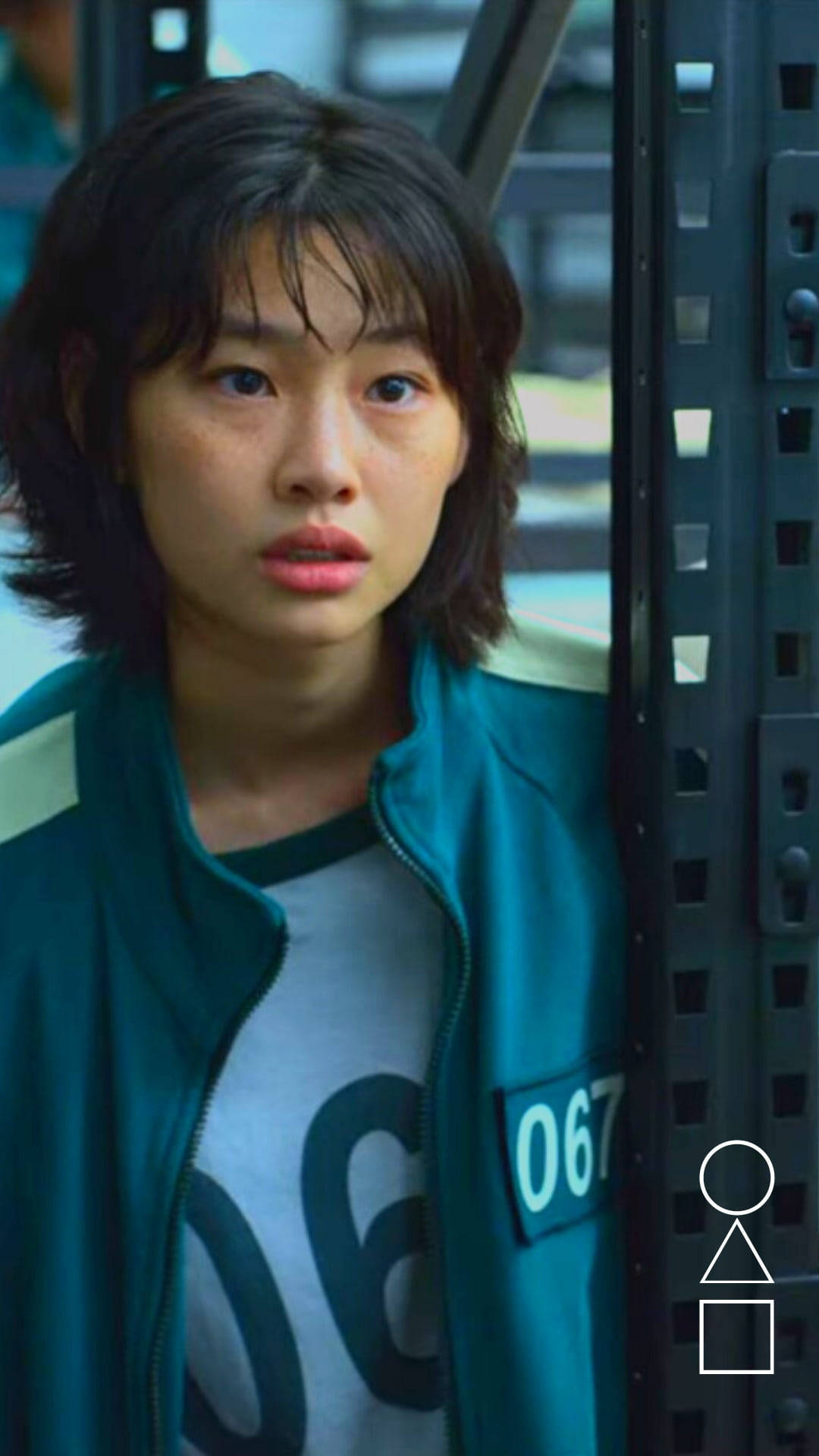 魷魚遊戲 067 號寢室對話 壁紙 — 一位身穿青綠色號碼外套的年輕女子，神情緊張地透過金屬格柵凝視著。
