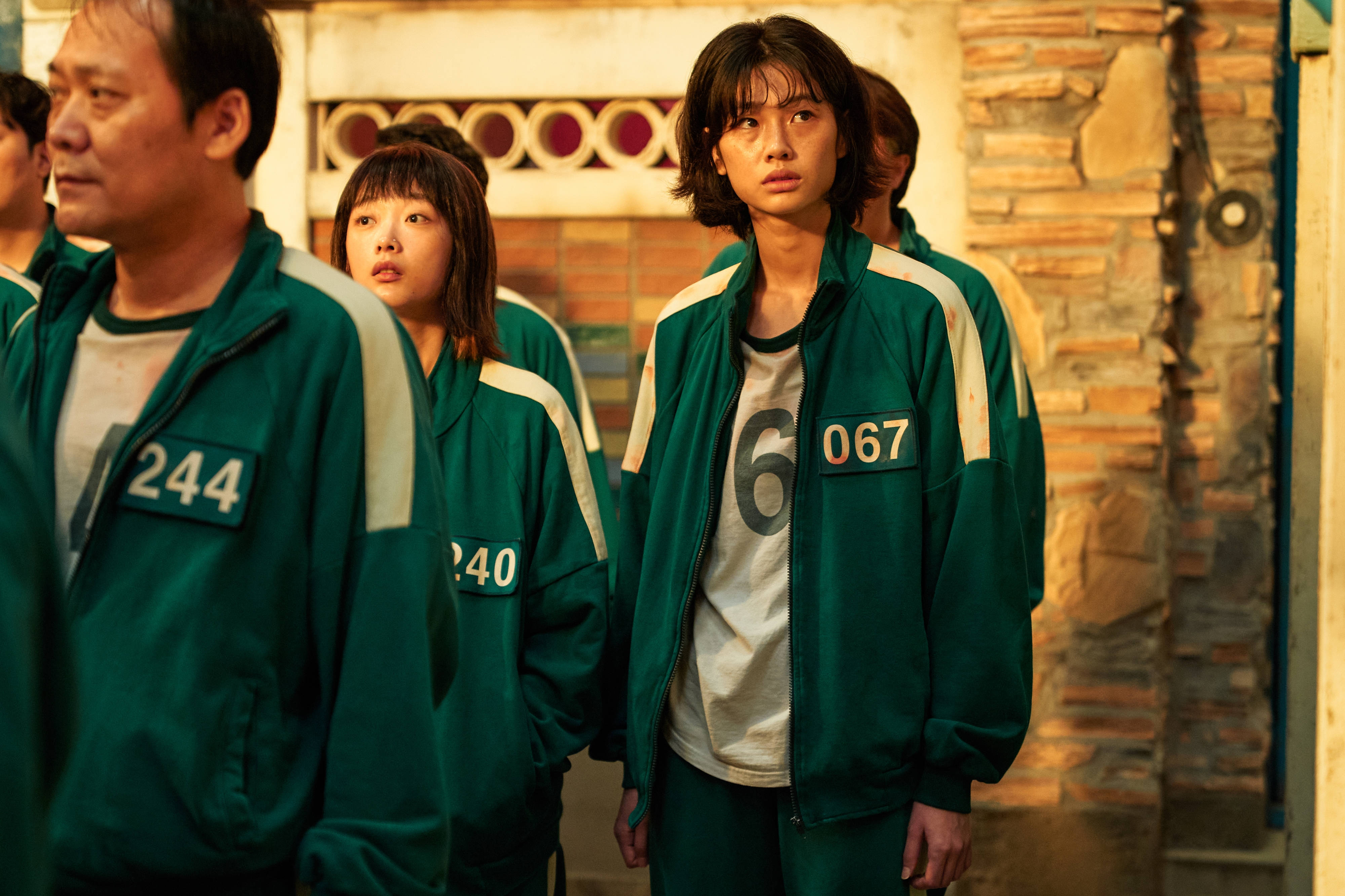 魷魚遊戲 067 號排隊取彈珠 壁紙 — 穿著綠色號碼運動服的參與者站在一條光線昏暗、有石牆和溫暖環境照明的巷子裡。