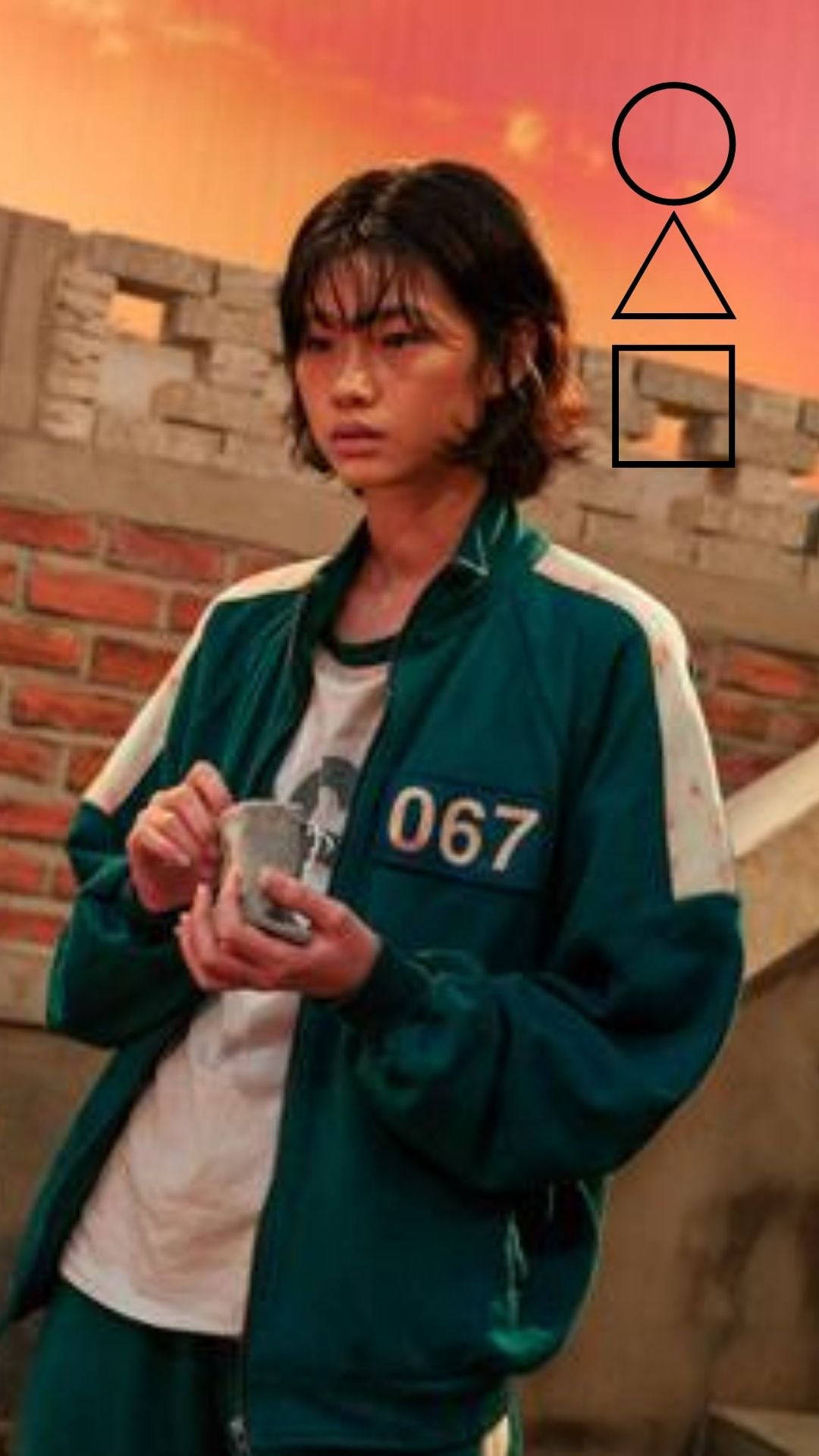魷魚遊戲 067 號握彈珠袋 壁紙 — 一個穿著綠色號碼外套的人，在火紅橙色的天空下，拿著杯子靠著磚牆。