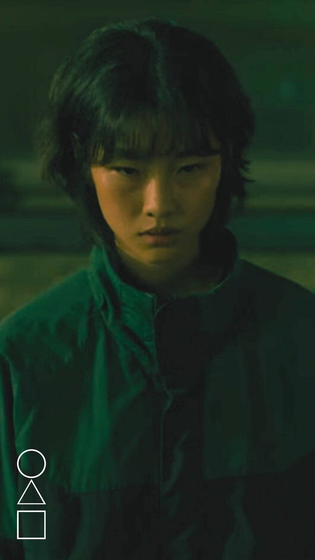 魷魚遊戲 067 號揭開黑暗之謎 壁紙 — 一位深色頭髮的年輕人在昏暗的綠色燈光下，穿著相配的綠色外套，專注地向前凝視。