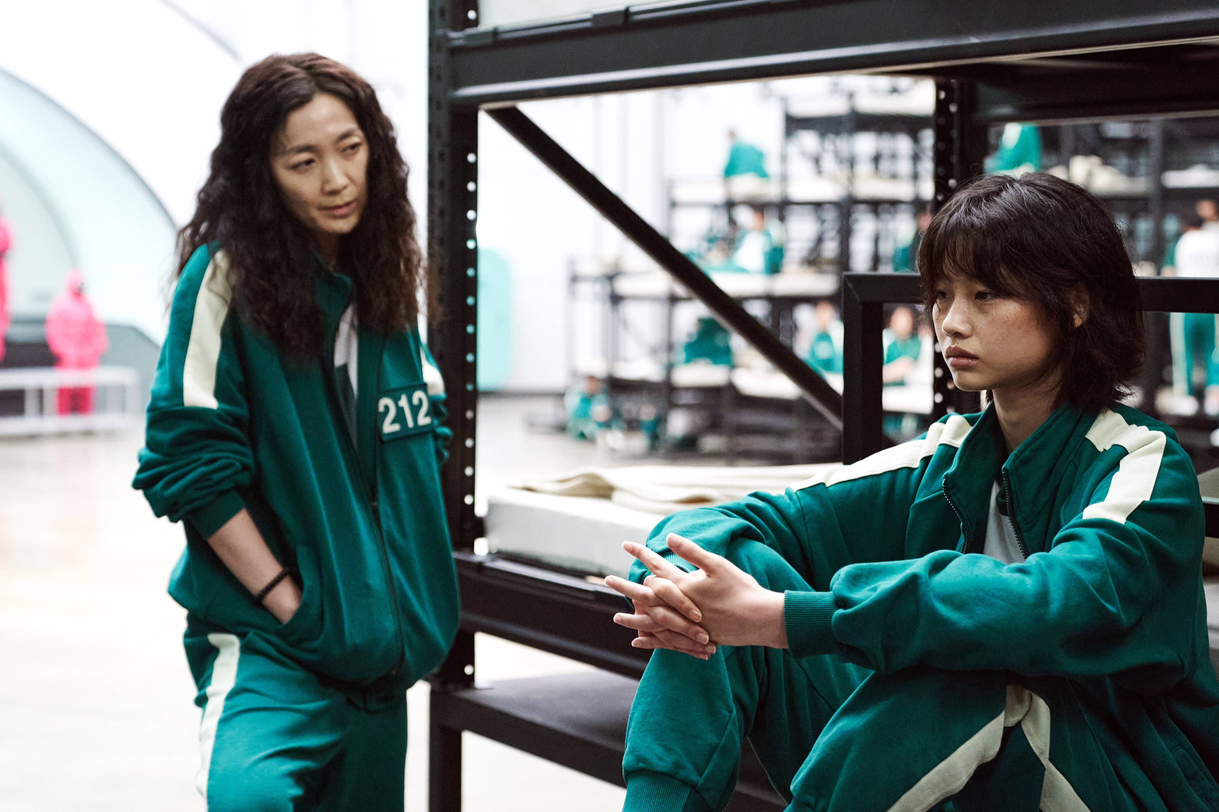 魷魚遊戲 067 號玩家 212 壁紙 — 兩名身穿綠色號碼運動服的女性坐在一個空曠的倉庫裡，背景有金屬架和其他球員。