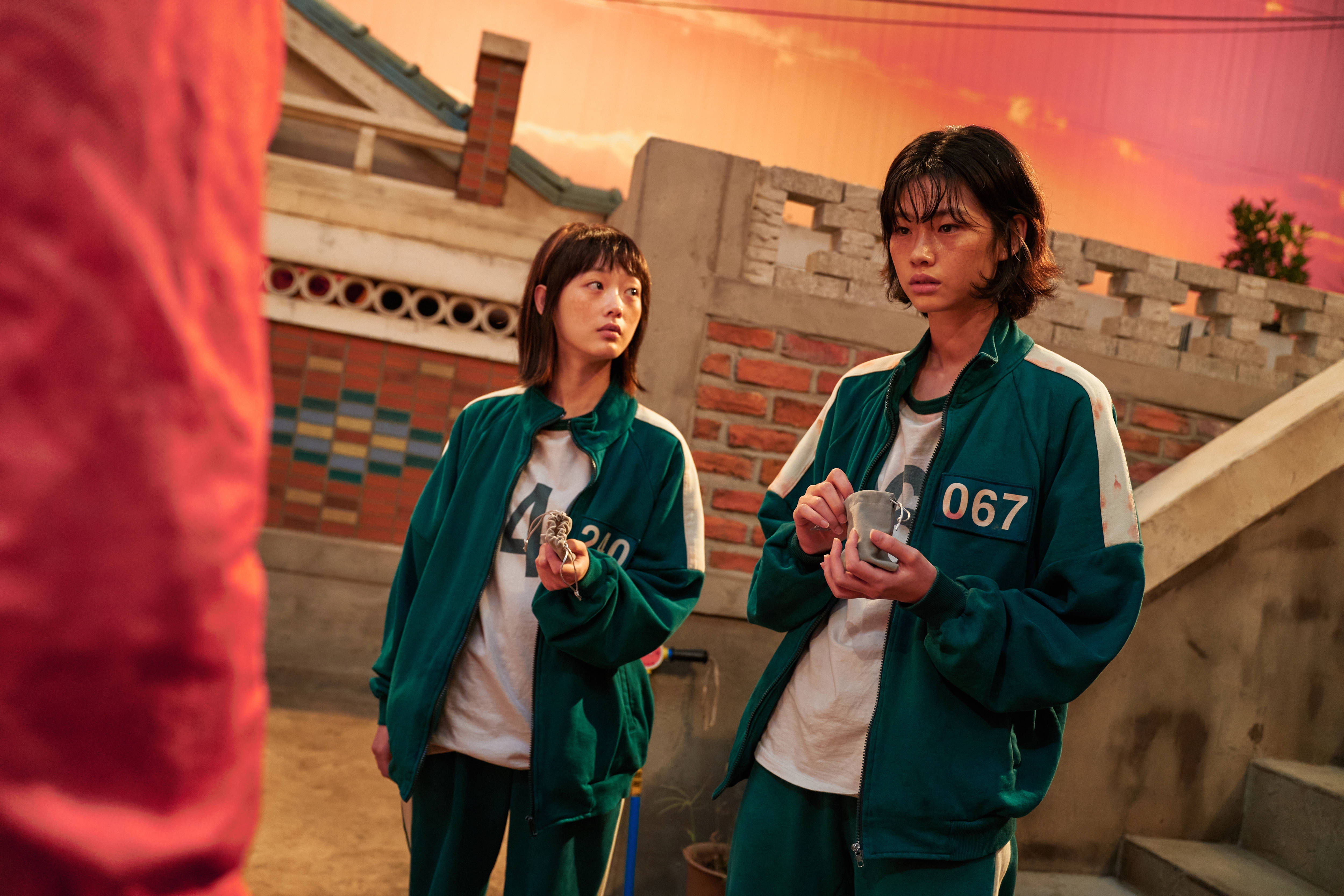 魷魚遊戲 067 配對賽 壁紙 — 兩名身穿綠色號碼運動服的球員在庭院中緊張地站着，夕陽將天空染成火熱的橙色和粉紅色。