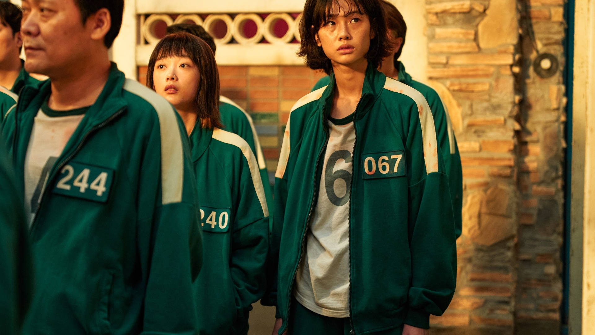 魷魚遊戲 067 高個子 壁紙 — 一群穿著綠色編號外套的人站在光線昏暗的石頭走廊中，表情焦慮。