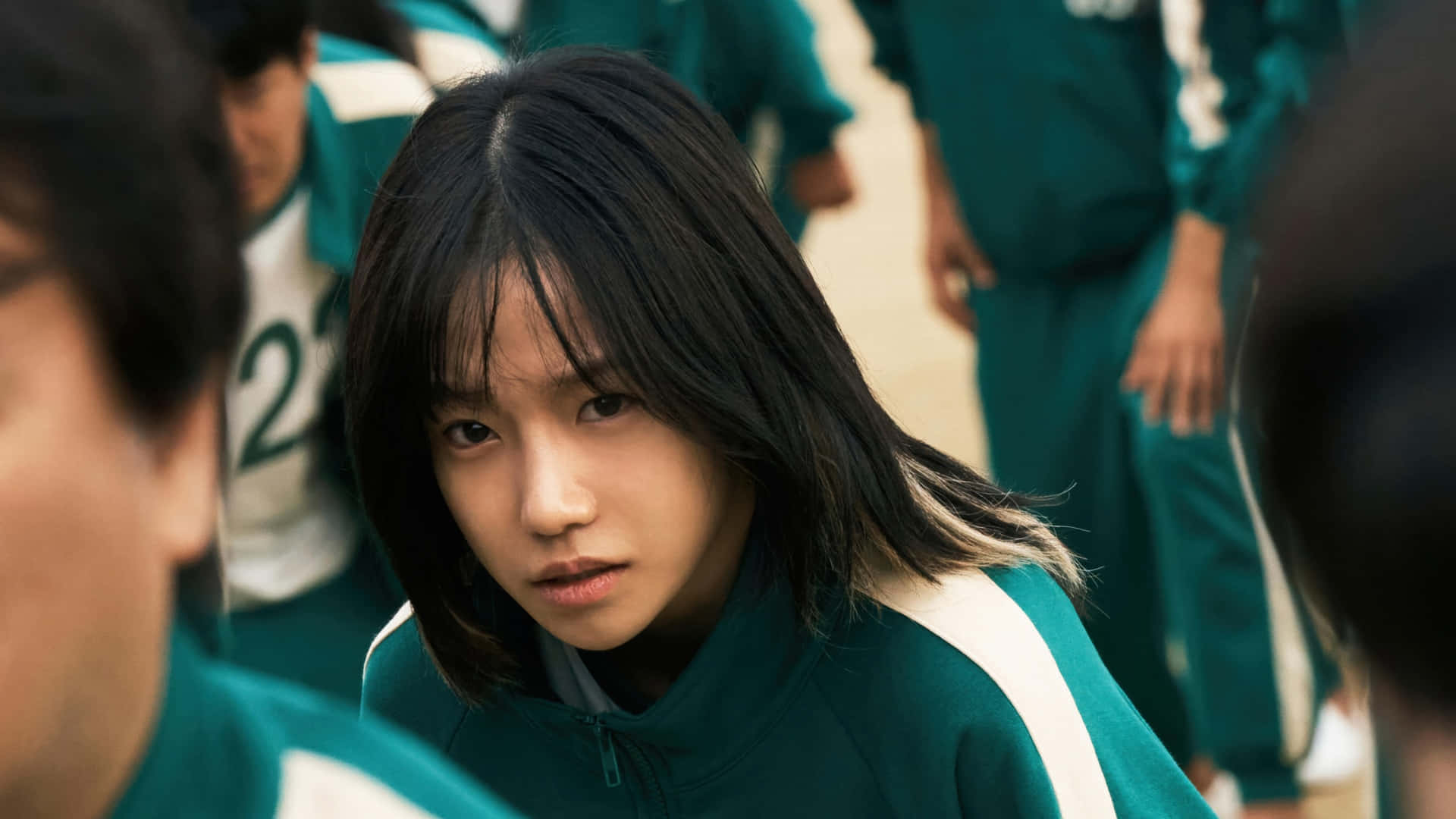 《魷魚遊戲》第二季預覽 壁紙 — 一位身穿綠色和白色運動服的年輕女性，在周圍穿著類似制服的人群中，神情緊張地凝視前方。