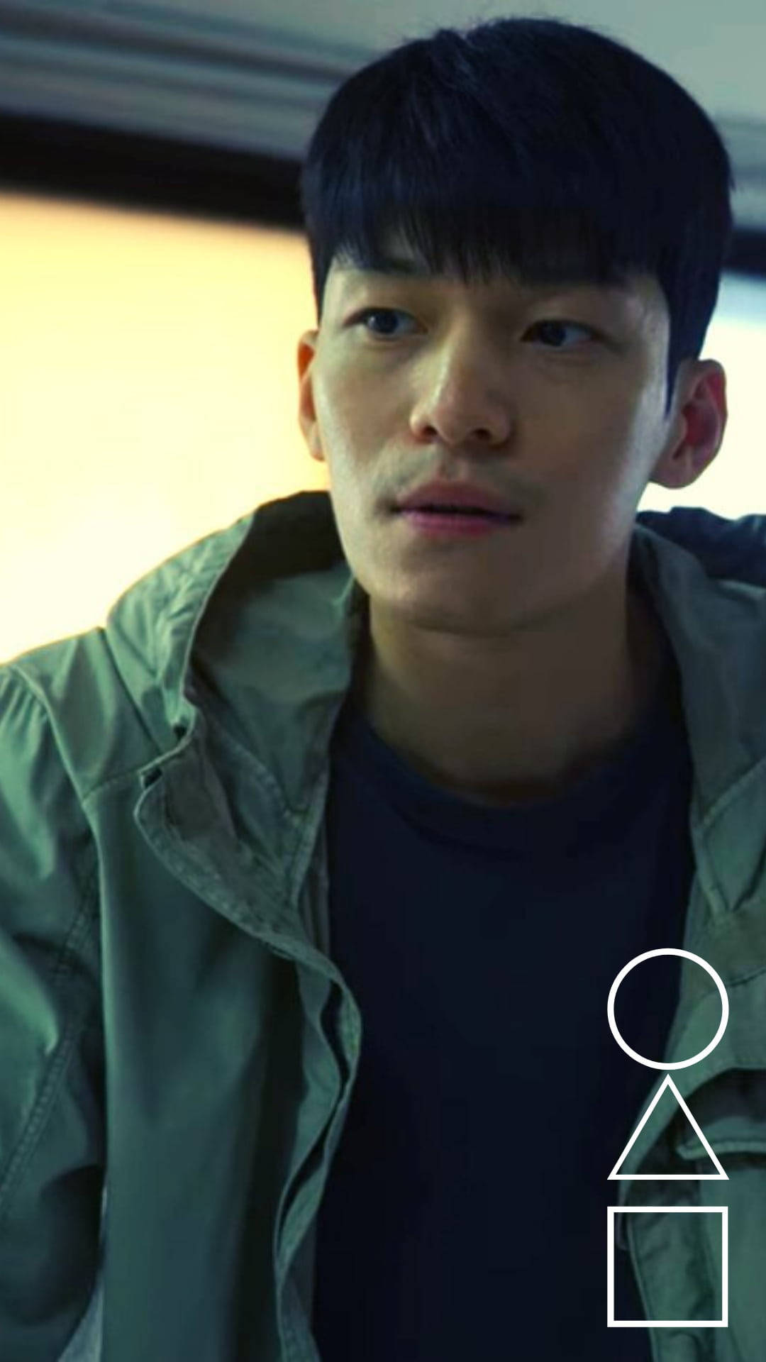 年輕男子穿著綠色外套，表情嚴肅地望向鏡頭外的室內場景。