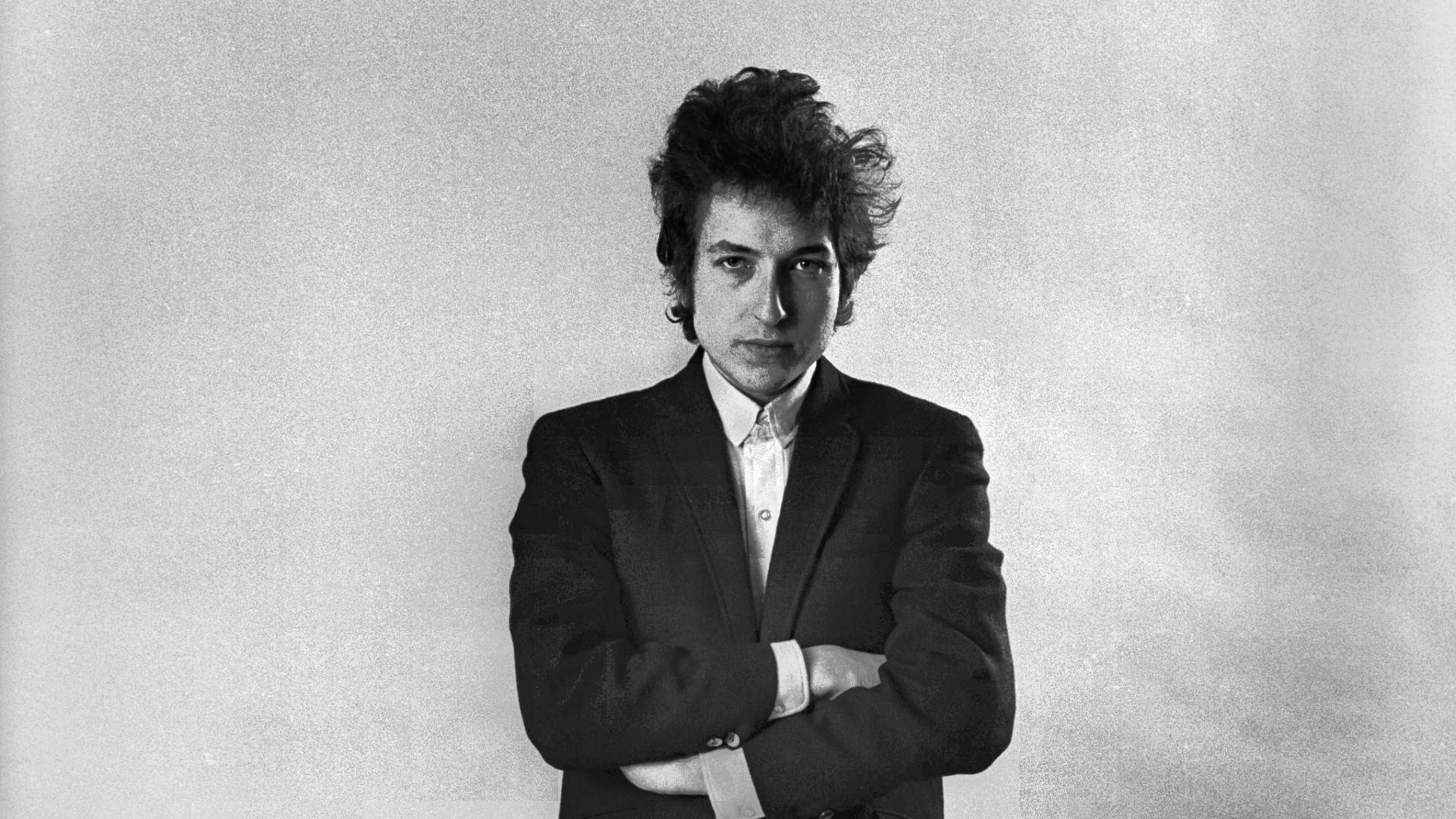 Bob Dylan 穿著黑色西裝交叉雙臂嘅工作室肖像。
