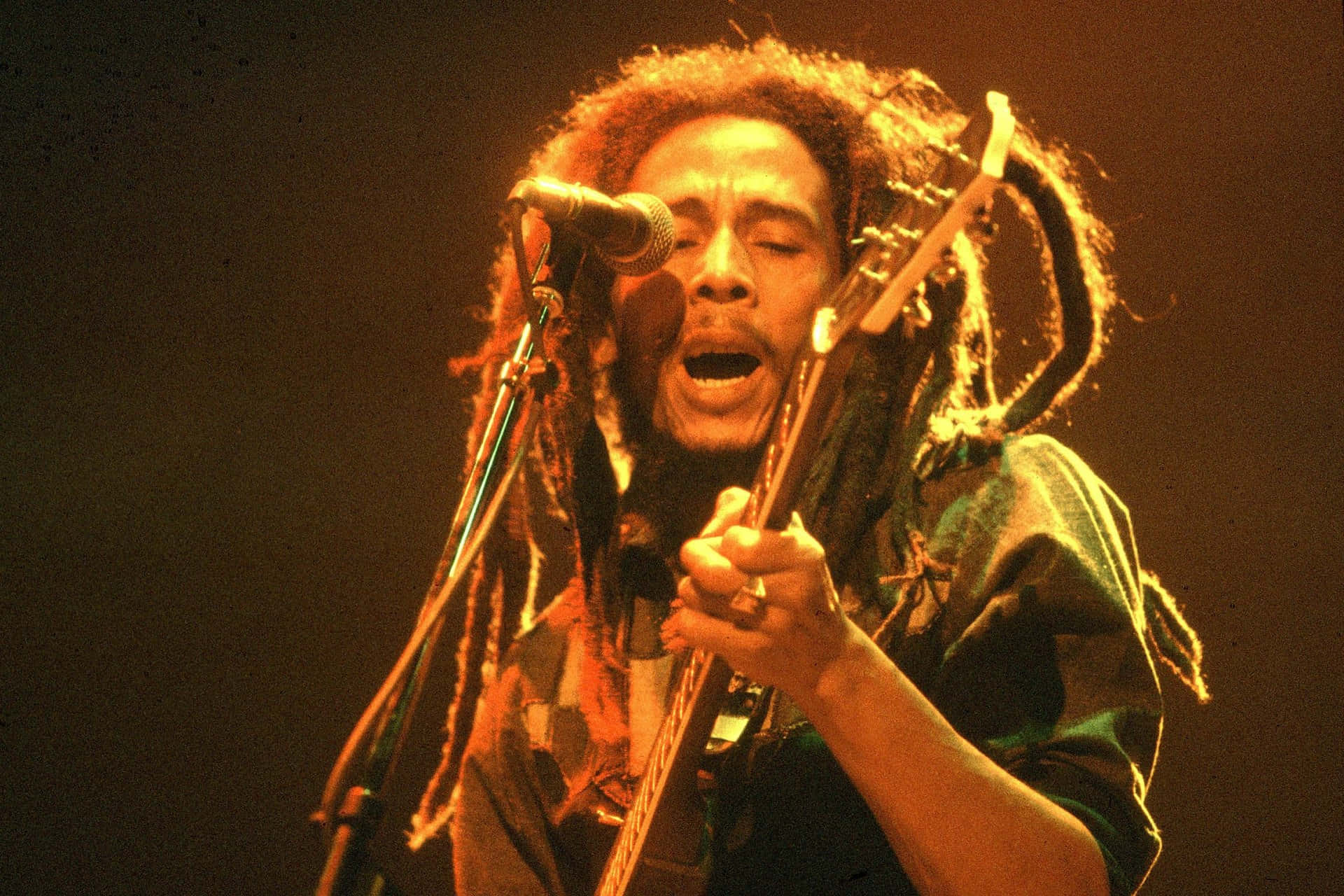 鮑勃·馬利 - 雷鬼傳奇 壁紙 — Bob Marley 在溫暖的舞台燈光下，熱情地對著咪高峰唱歌，同時彈奏結他。
