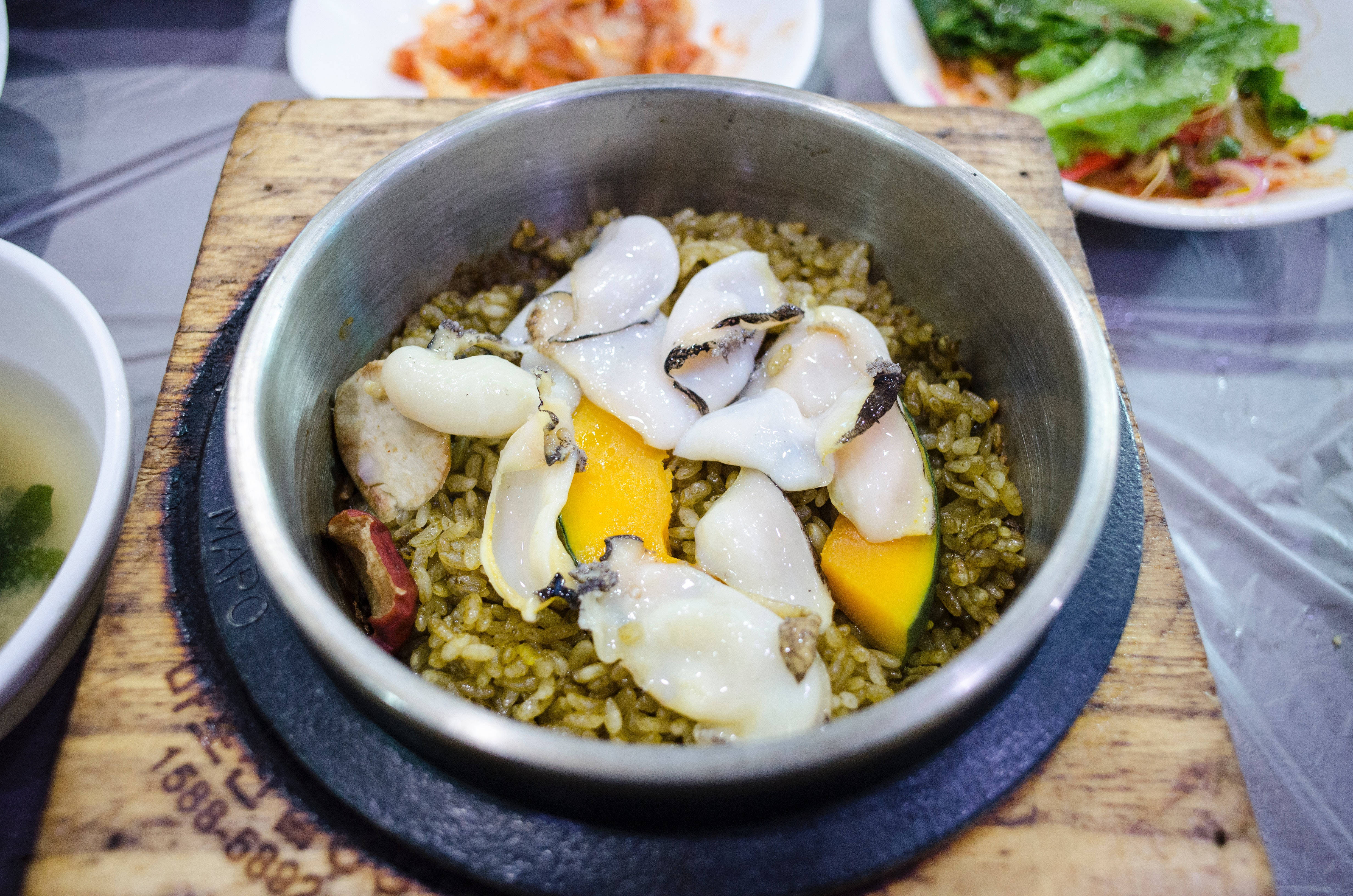 鮑魚韓式熱鍋飯 壁紙 — 一個冒煙的石鍋，盛載著韓國海鮮飯，配以生蠔、芒果和青菜，放在木板上。