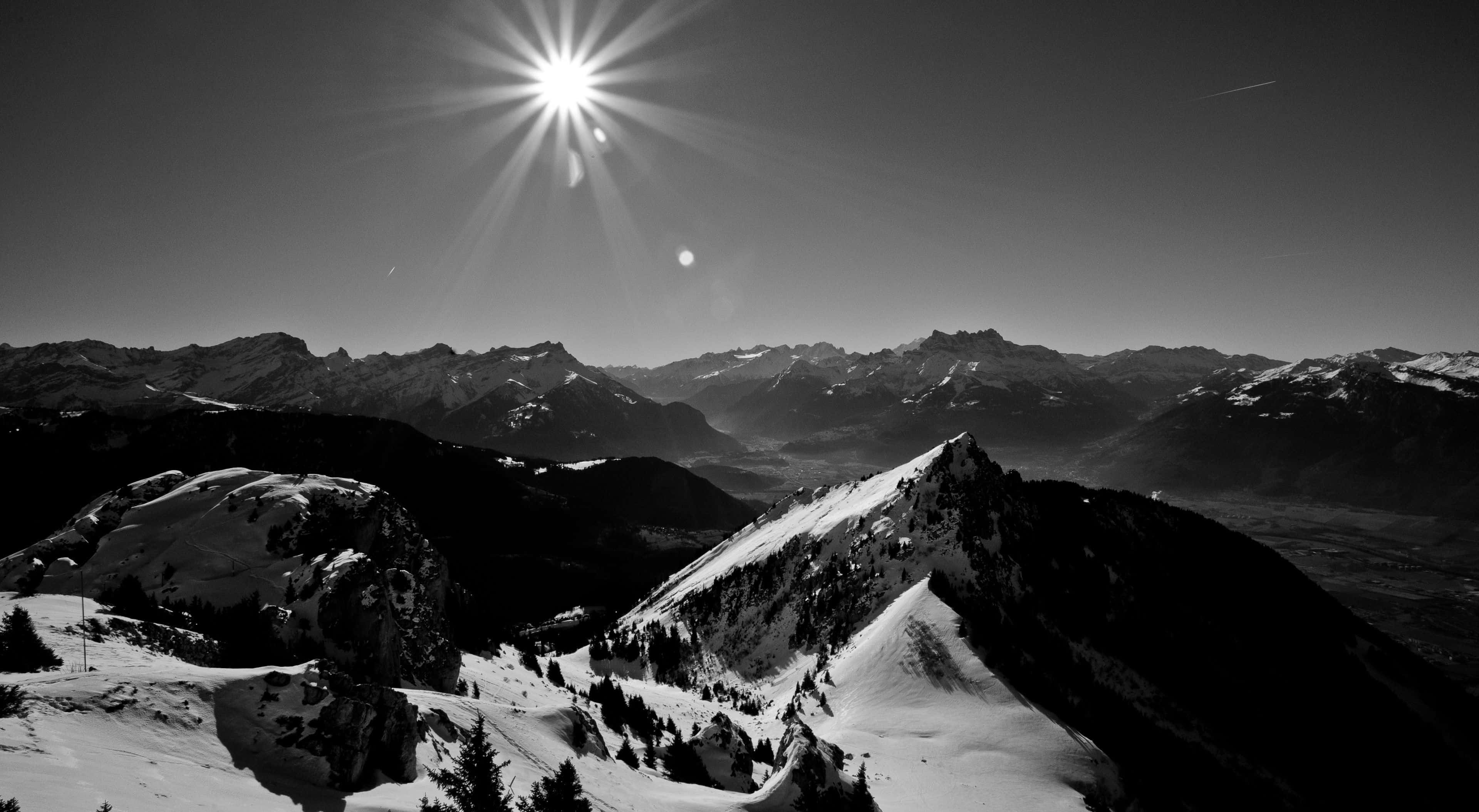 Black And White Landscape 桌布 壁紙 — 鮮明的黑白山景，一輪光芒四射的太陽在積雪的山峰和深邃的山谷上投下銳利的光芒。