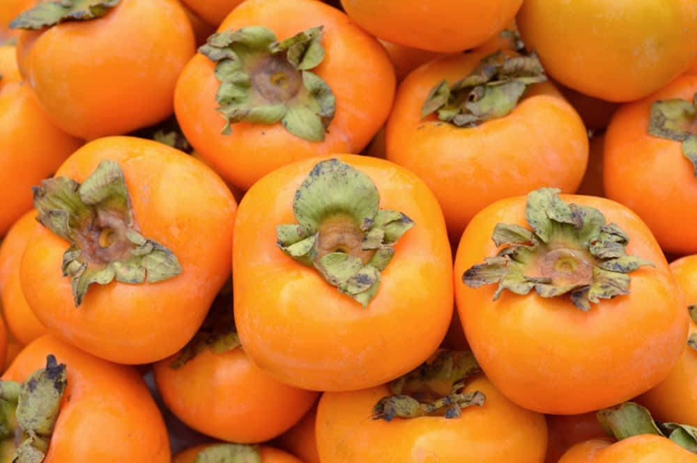 Winter Fruits 桌布 壁紙 — 鮮橙色柿子和綠色葉莖堆疊在一起的近距離特寫。