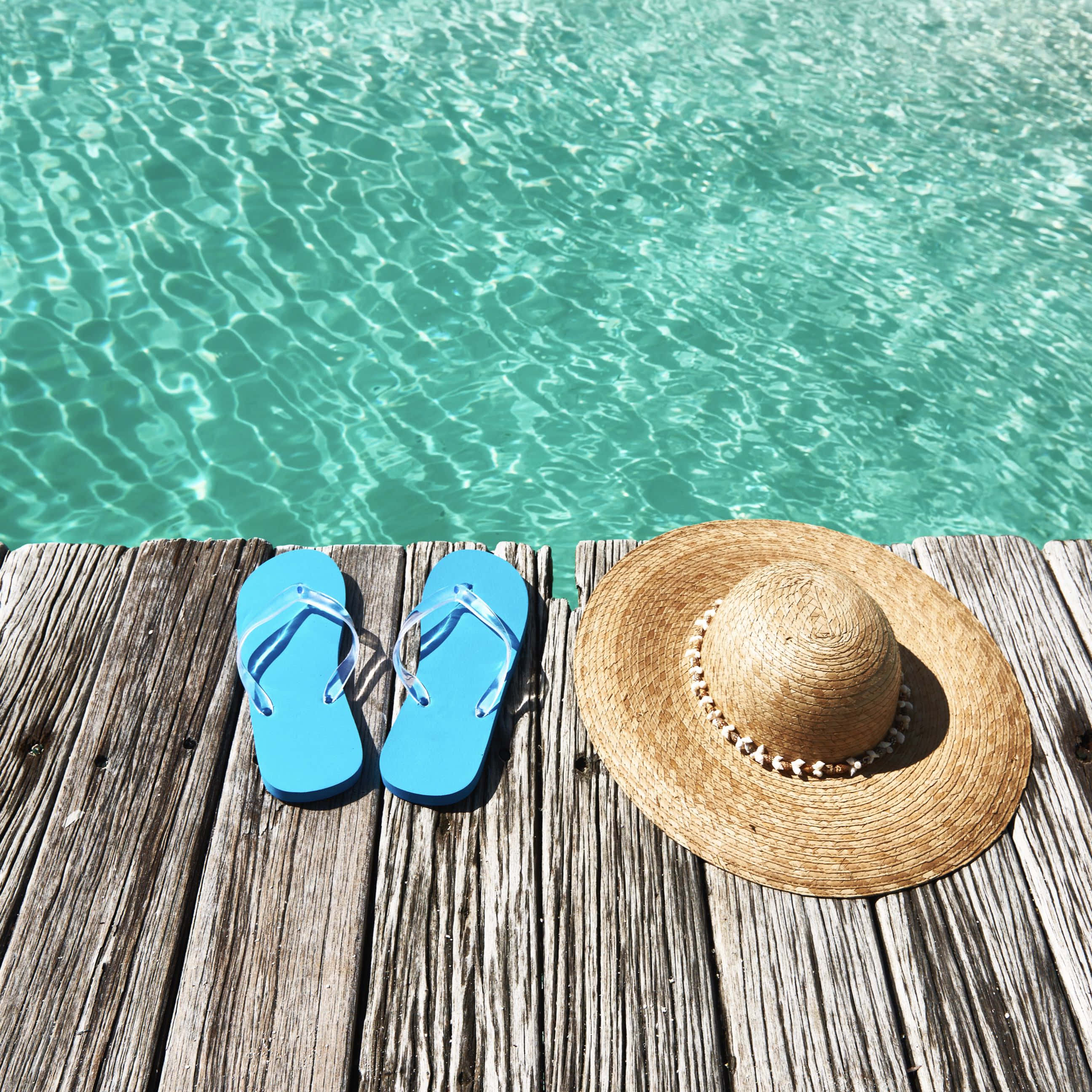Beach Sandals 桌布 壁紙 — 鮮藍色人字拖和草帽放在木製碼頭上，旁邊是閃閃發光的碧綠色泳池水。