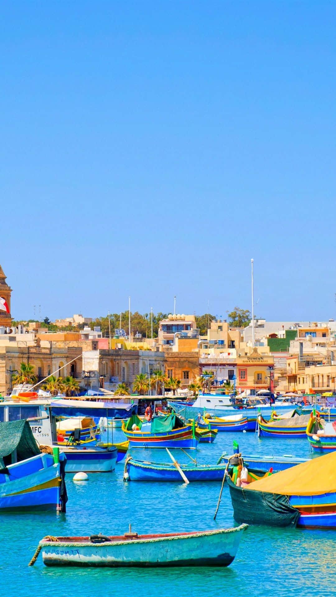 Malta 桌布 壁紙 — 鮮藍色漁船在清澈天空下的碧綠海水中搖曳，襯托著陽光普照的地中海建築。