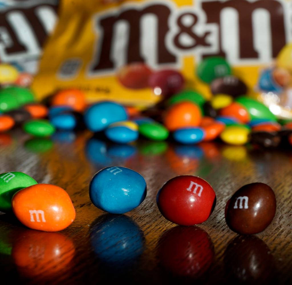 Chocolate 桌布 壁紙 — 鮮豔的M&M's糖果在前景，後面有模糊的黃色袋子在反光的木質表面上。