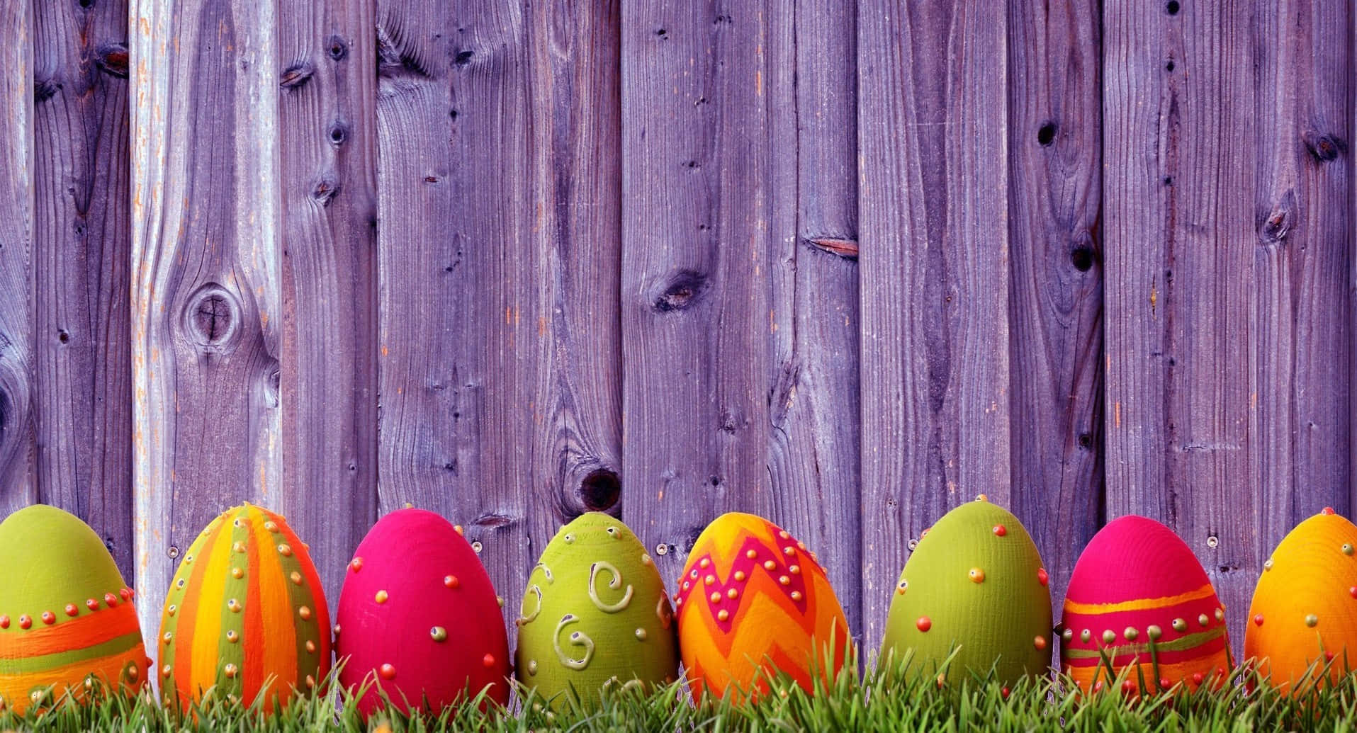 Eggs 桌布 壁紙 — 鮮豔的手繪復活節彩蛋帶有閃亮圖案，放置在綠色草地上，背景是風化的紫色木柵欄。