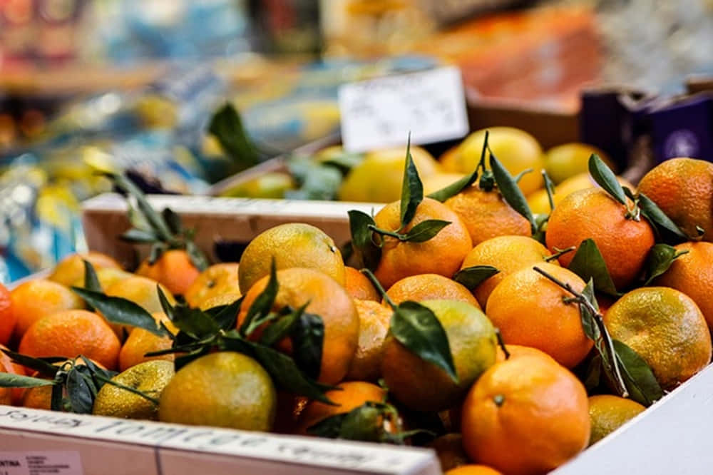 Winter Fruits 桌布 壁紙 — 鮮豔的橙子連同綠葉堆疊在紙箱中，位於熱鬧的市場攤位。