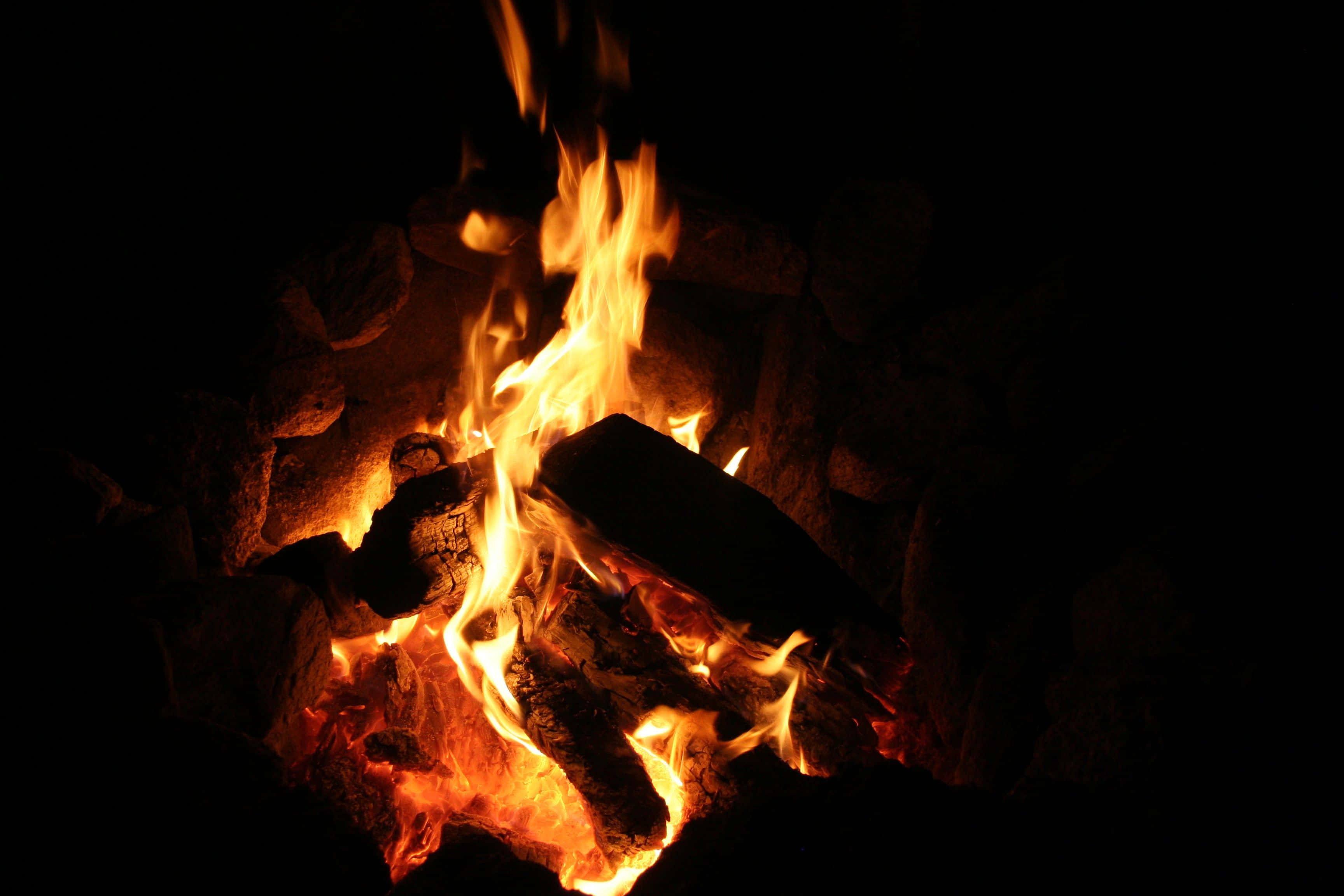 Campfire 桌布 壁紙 — 鮮豔的橙色火焰在漆黑背景中，圍繞著嵌在深色岩石中的發光餘燼跳躍舞動。