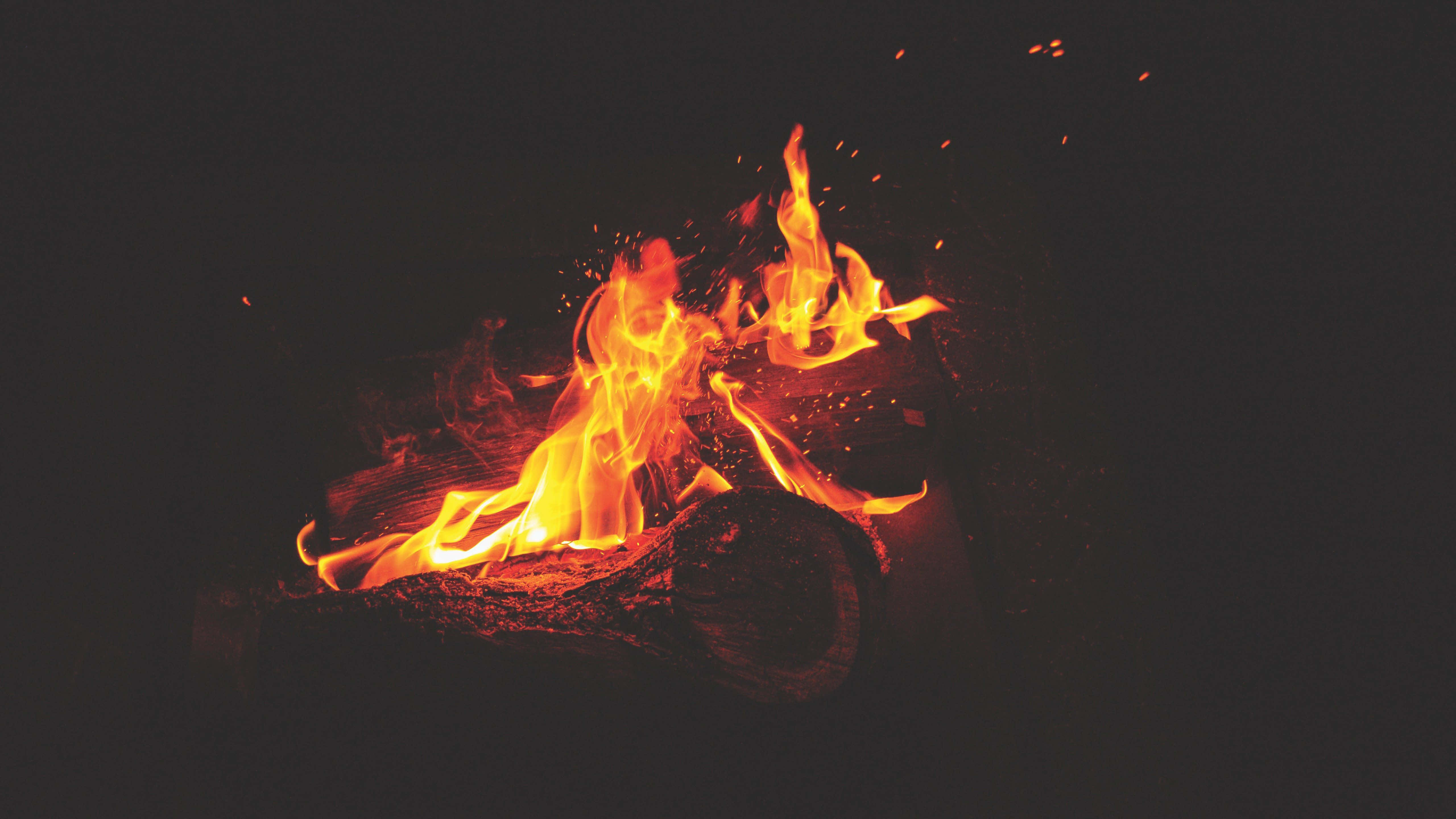 Campfire 桌布 壁紙 — 鮮豔的橙色火焰在燒焦的木頭上跳躍並發出劈啪聲，背景是深邃的黑色夜空，發光的餘燼向上飄浮。