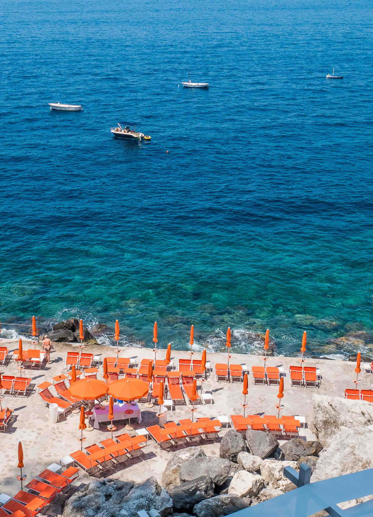 Italian Beach 桌布 壁紙 — 鮮豔的橙色躺椅和遮陽傘排列在岩石懸崖海灘上，俯瞰著碧綠的海水，遠處有小船漂流。