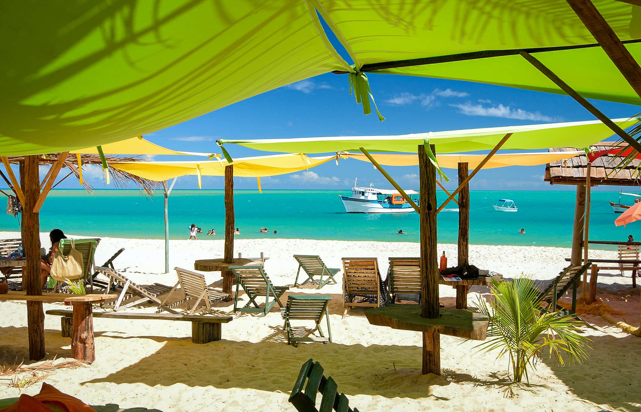 Beautiful Beach 桌布 壁紙 — 鮮豔的檸檬綠和黃色遮陽篷陰影覆蓋在木製躺椅上，位於純淨的白色沙灘上，背景是碧綠的海水和船隻，天空清澈湛藍。
