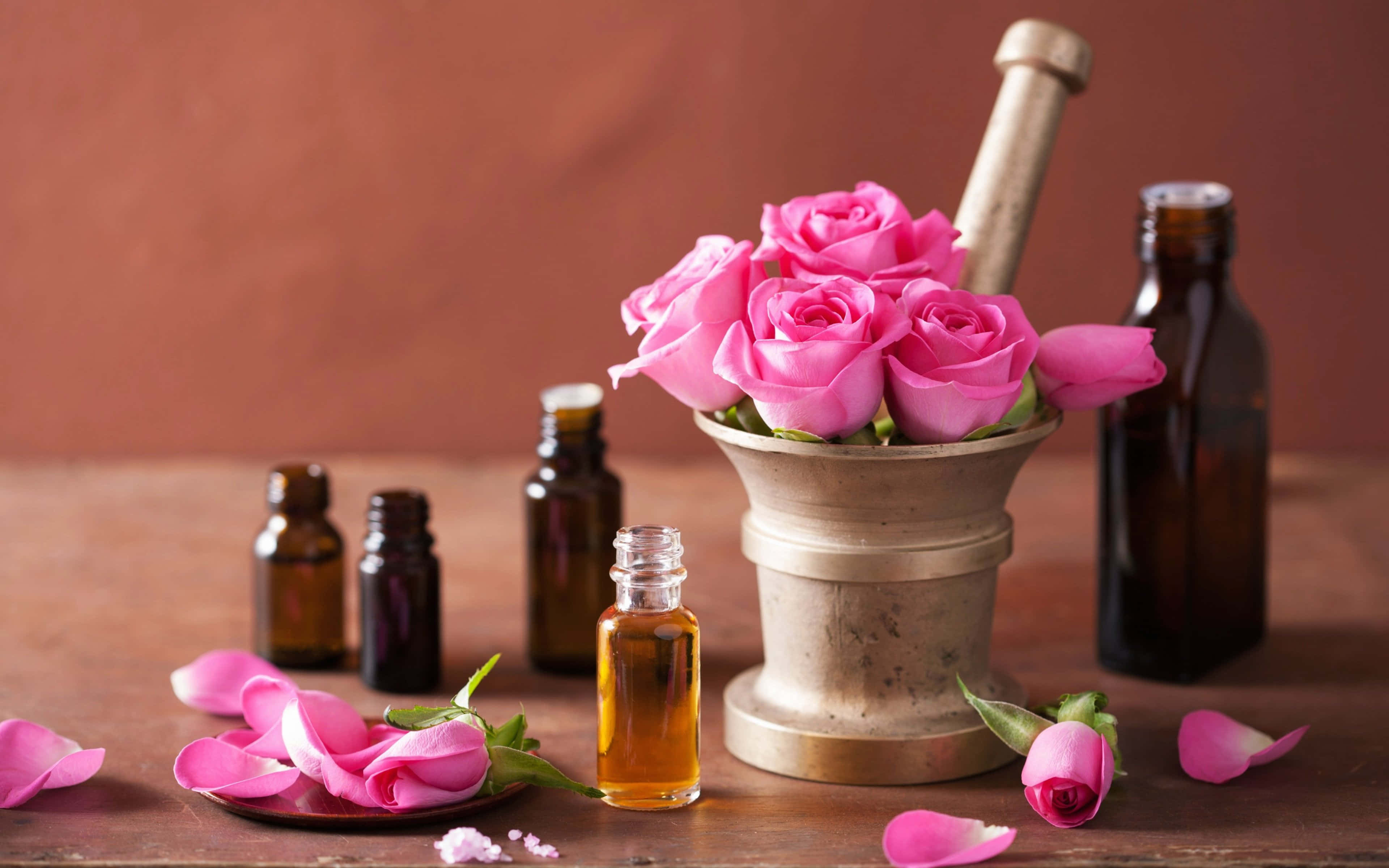 Floral Fragrance 桌布 壁紙 — 鮮豔的粉紅玫瑰在黃銅研缽中盛開，旁邊是琥珀色的精油瓶，放在質樸的木桌上。