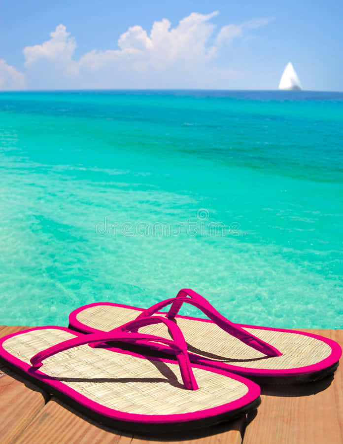Beach Sandals 桌布 壁紙 — 鮮豔的粉紅色人字拖放在木甲板上，俯瞰著帶有帆船和藍天的碧綠海洋。