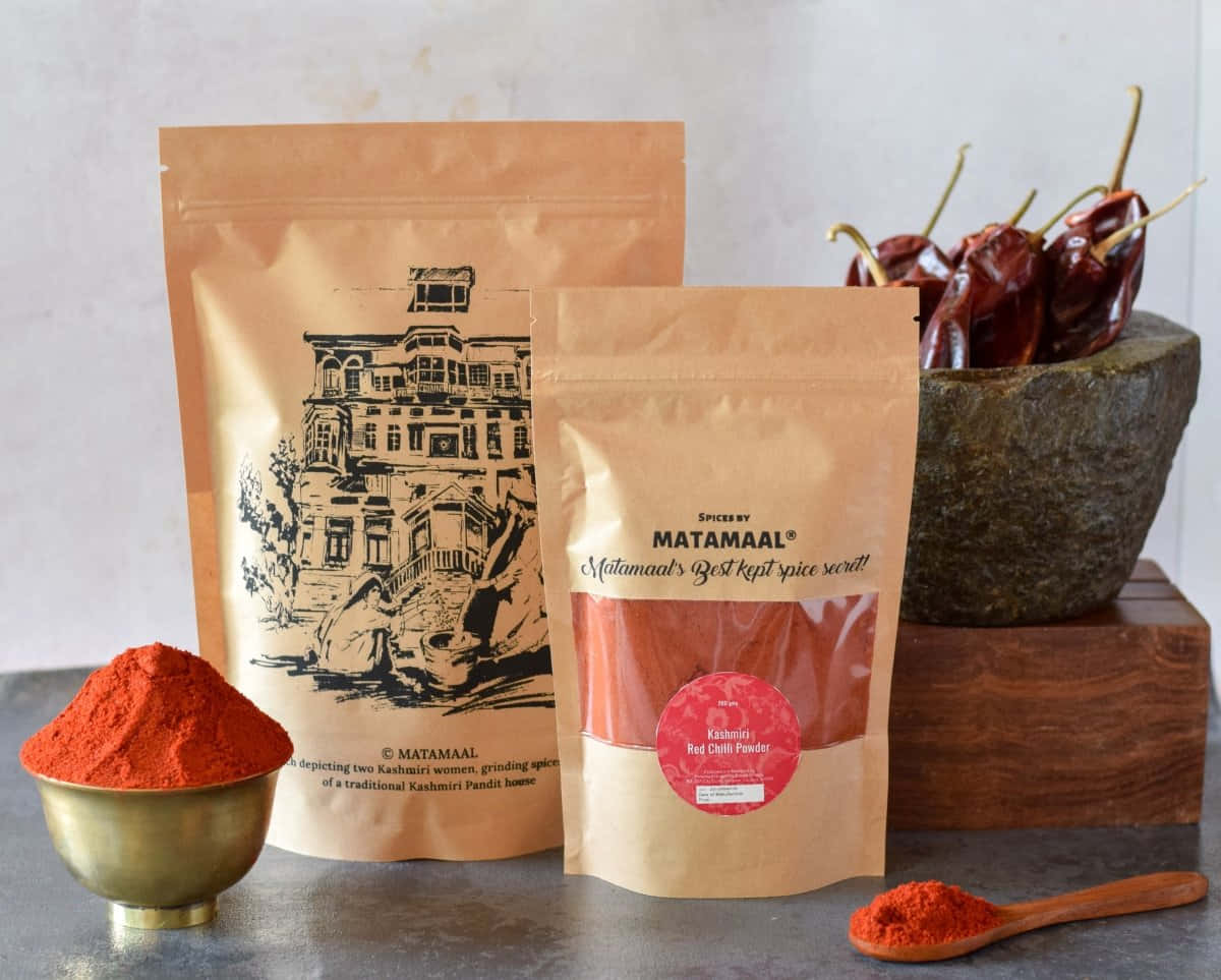 Red Chili Powder 桌布 壁紙 — 鮮豔的紅色克什米爾辣椒粉放在黃銅碗中，旁邊有品牌香料袋和石臼中的乾辣椒。